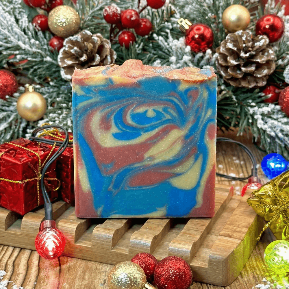 Swirled bar soap with marbled blue, gold, and rose colors on a wooden soap dish, surrounded by festive Christmas decorations including ornaments, pine branches, and wrapped gifts.