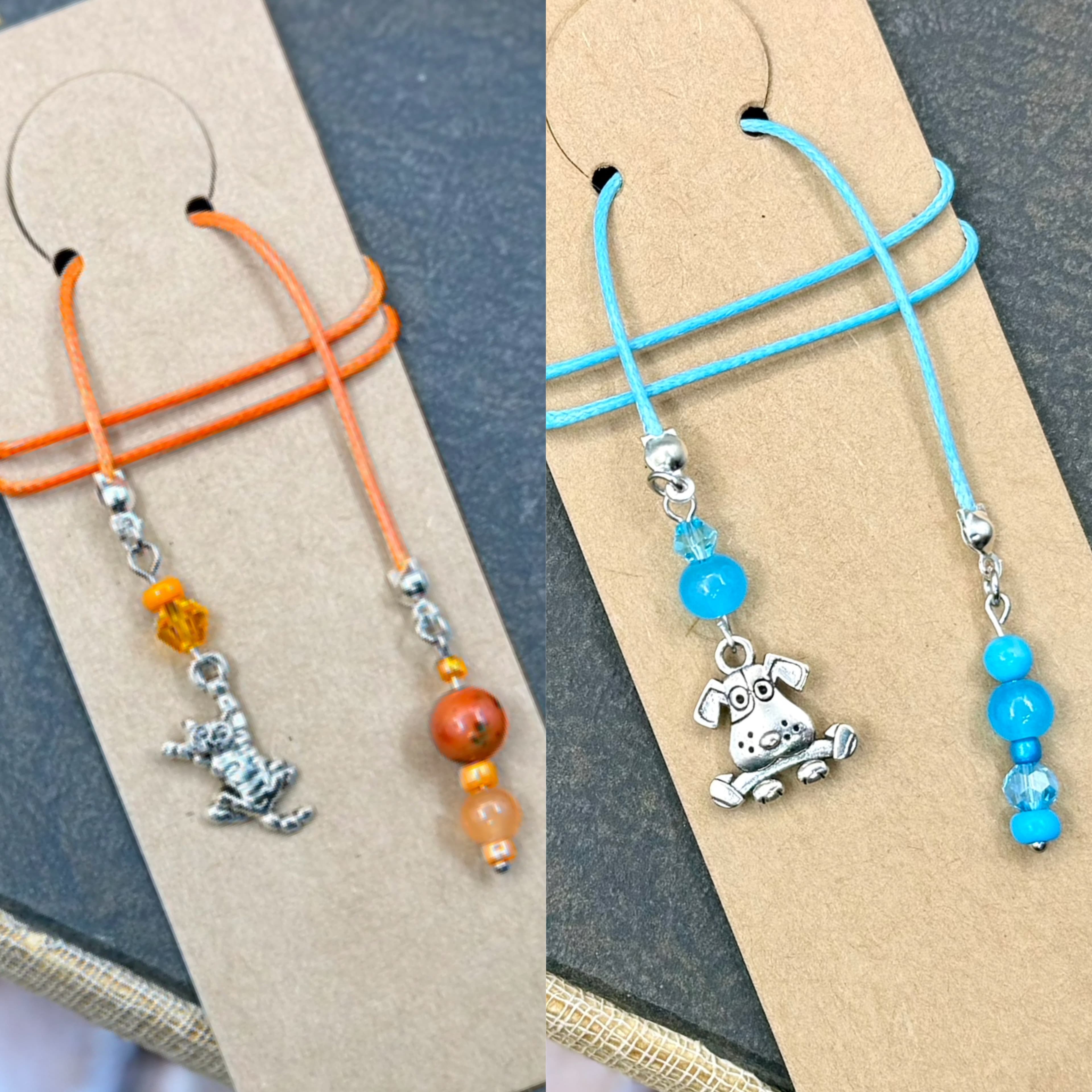 Two handmade bookmarks displayed on cardstock, one with orange cord and cat charm with orange beads, the other with blue cord and dog charm with blue beads.
