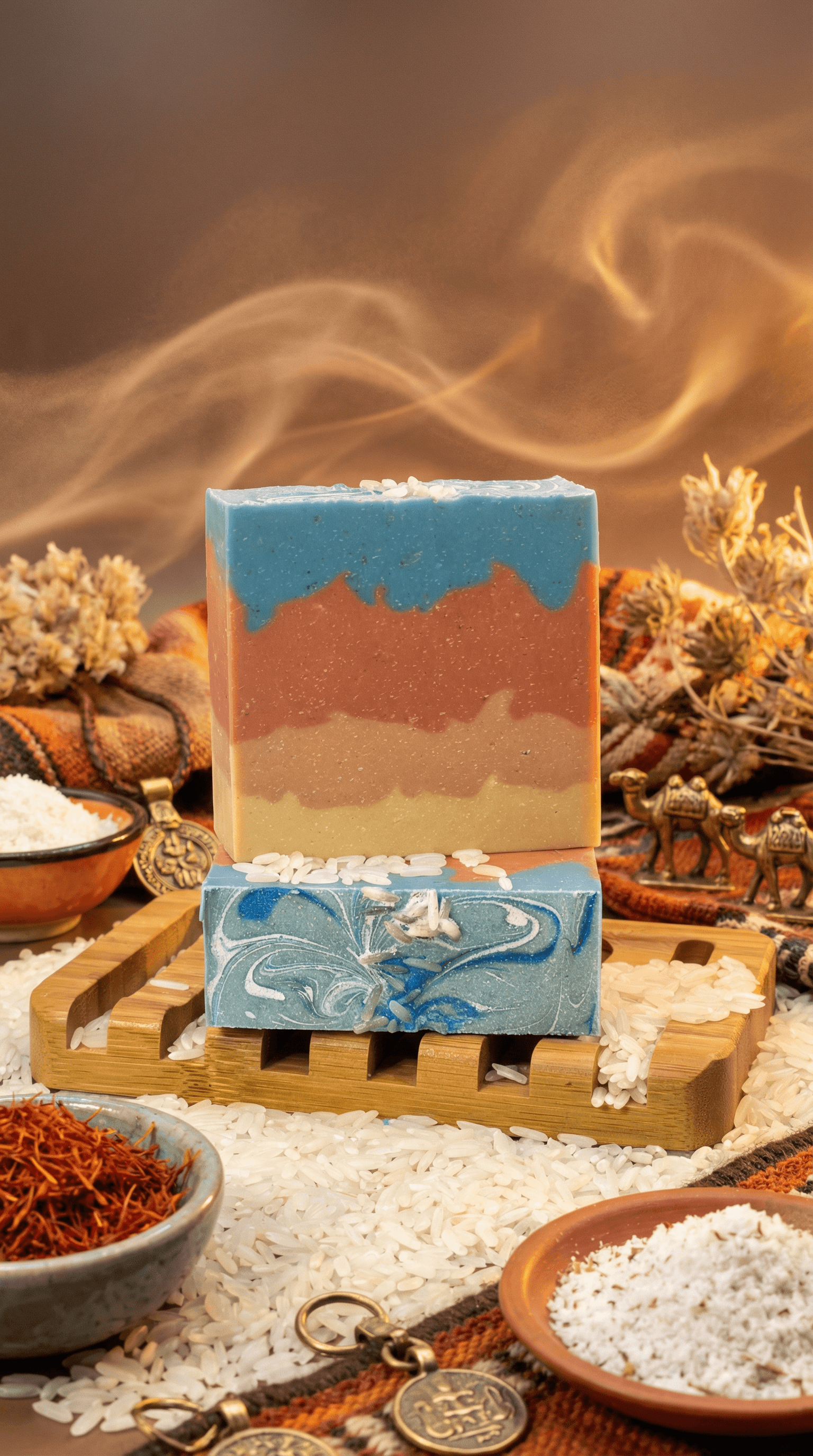 Two handcrafted soap bars stacked on a wooden soap dish surrounded by dried botanicals, sea salt, and saffron. Top bar features desert sunset gradient layers in blue, rust, and tan.