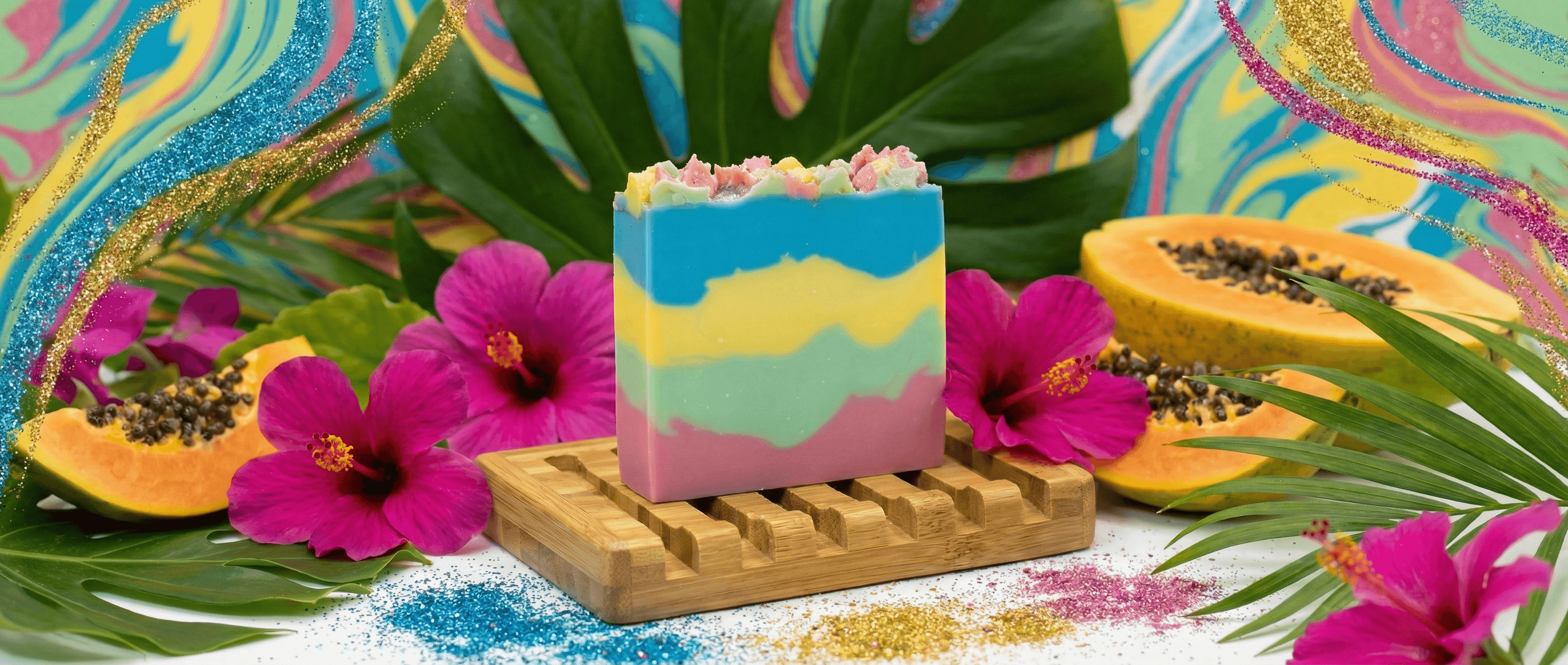 Handcrafted soap bar with horizontal wavy stripes in blue, yellow, mint green, and pink, topped with pastel cereal pieces. Displayed on bamboo dish surrounded by tropical leaves, hibiscus flowers, and papaya.