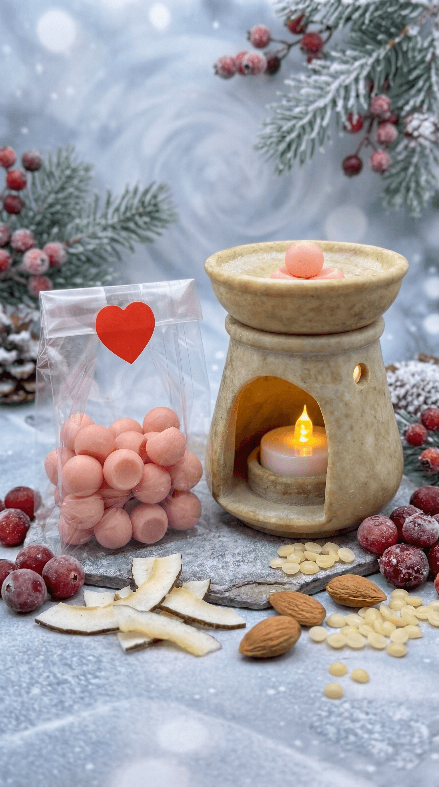 Beige ceramic wax warmer with lit tea light beside clear bag of peachy-pink wax melt spheres with red heart sticker. Winter styling with frosted evergreen, cranberries, and coconut chips.
