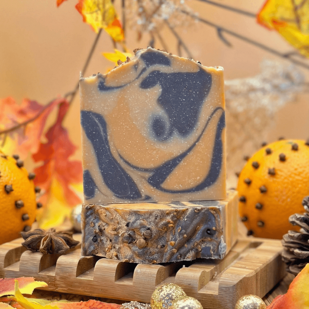 Two stacked bars of handcrafted soap with navy blue swirls on tan base, displayed on wooden soap dish. Bottom bar shows embedded botanicals including cloves and orange peel, surrounded by clove-studded oranges and autumn leaves.