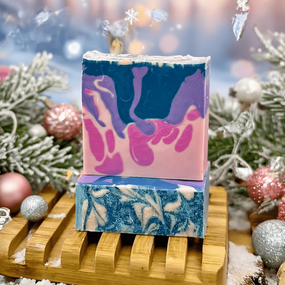 Two stacked handmade soap bars on wooden rack surrounded by holiday decor. Top bar features pink and teal swirled layers; bottom bar shows cream and teal marbling.