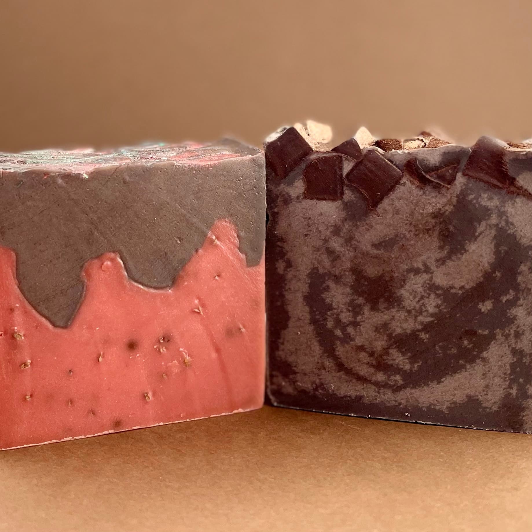 Two handcrafted soap bars side by side: one coral-pink with gray wavy top layer, one charcoal gray with chocolate chunk pieces on top. Both bars show textured surfaces against tan background.