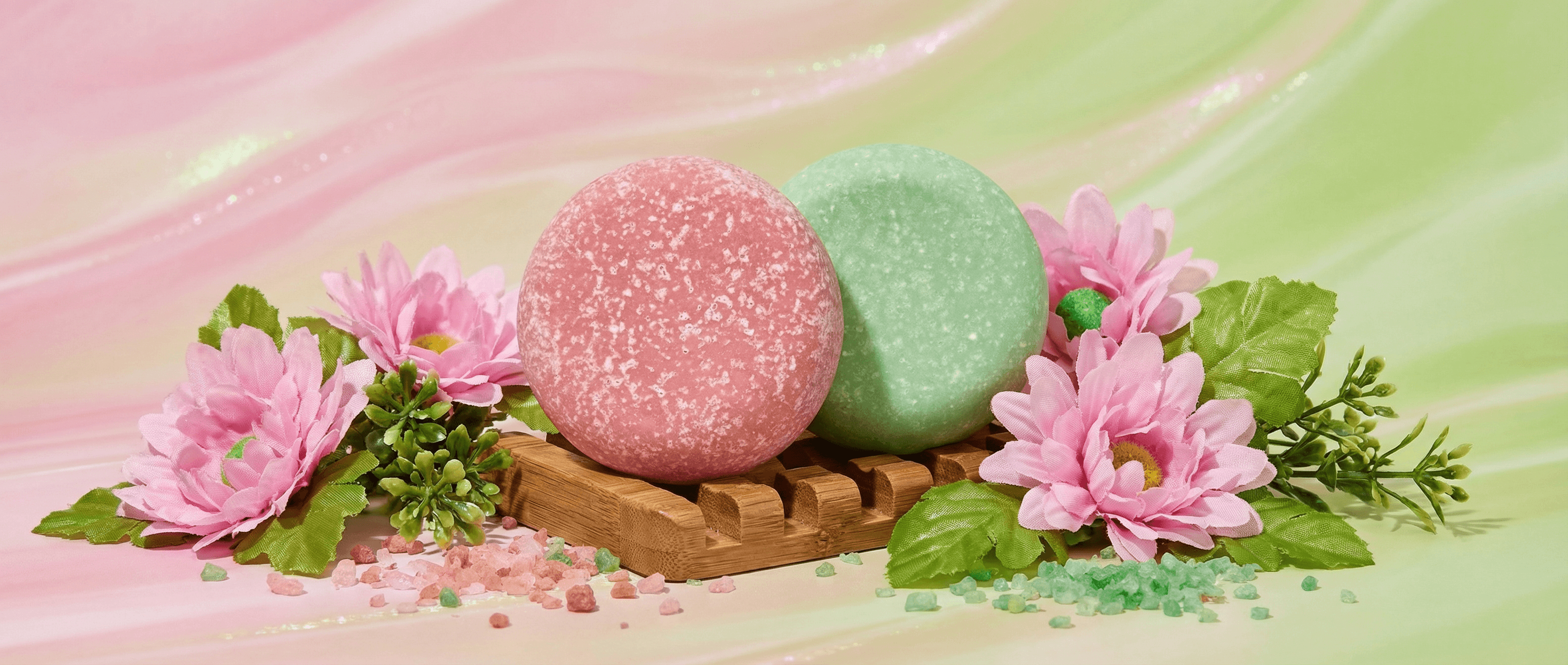 Two round sea salt scrub bars in pink and green with crystalline texture, displayed on a wooden soap dish with pink flowers and scattered salt crystals on a pastel background.