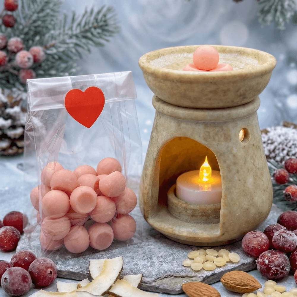 Beige ceramic oil warmer with lit tea light next to clear bag of peach-colored wax melt balls with red heart label. Styled with frosted cranberries, pine branches, and nuts.