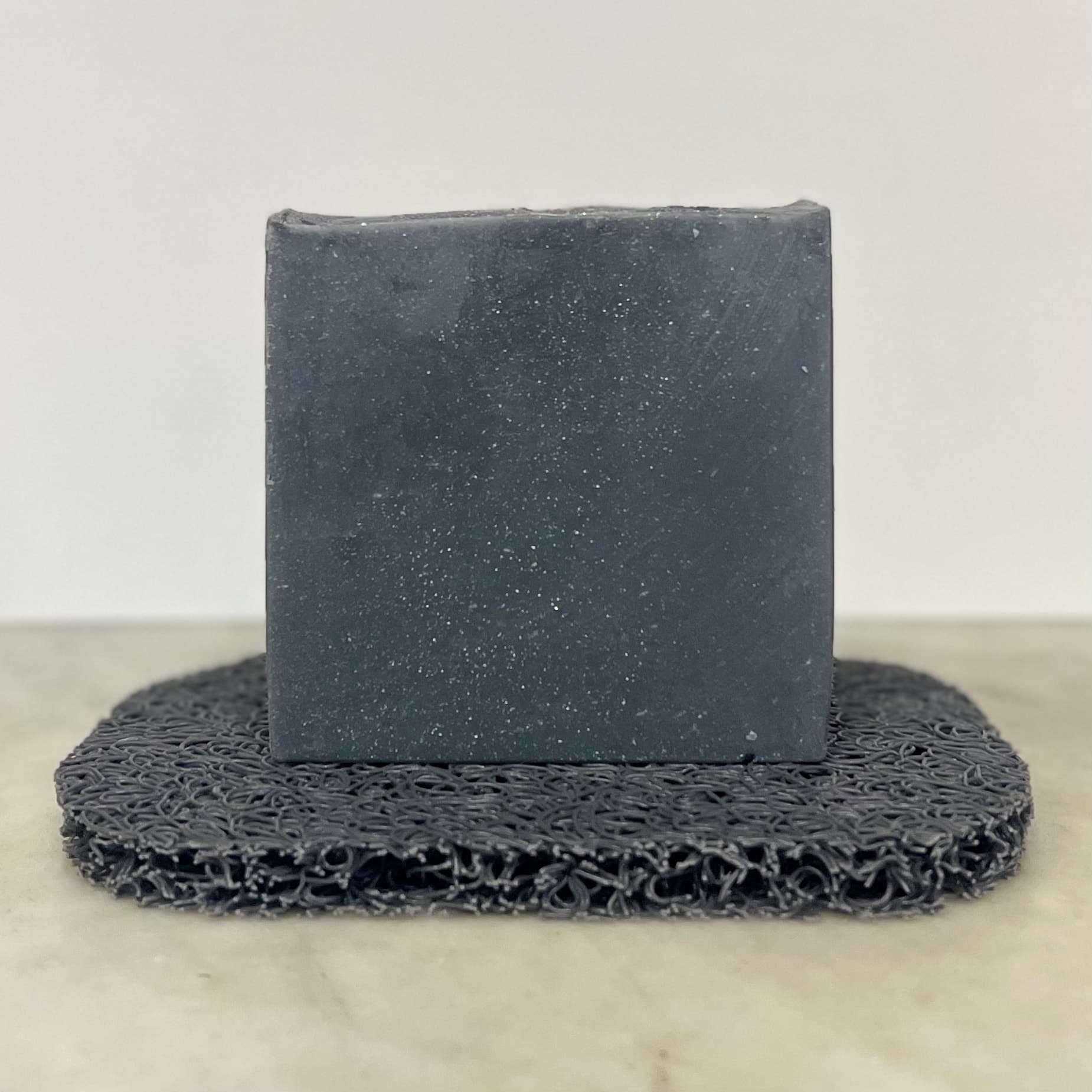 Dark gray-blue bar with fine white speckles throughout, sitting on a matching gray textured soap saver mat against a neutral background.