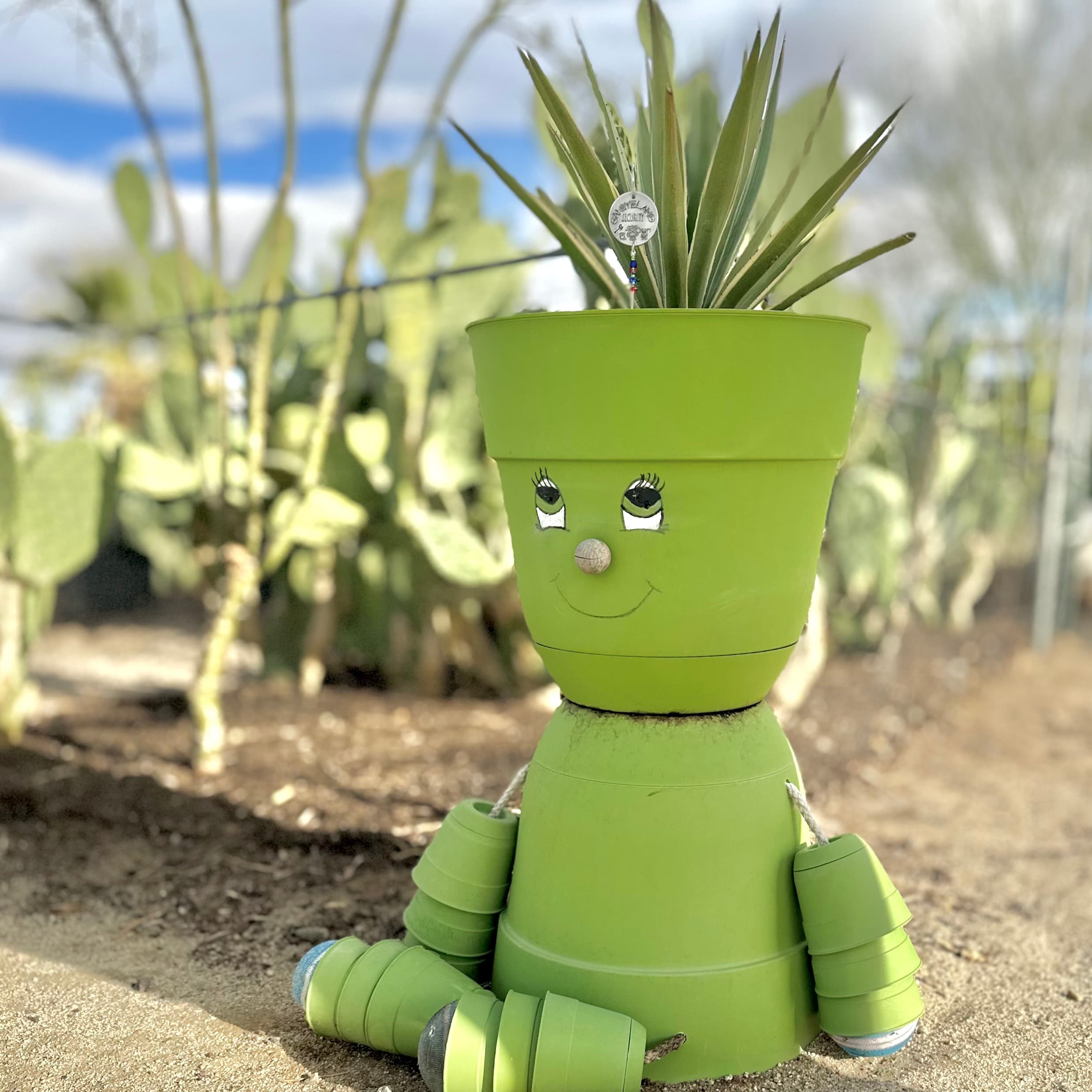 Lime green terracotta pot planter person with painted smiling face, seated in desert landscape surrounded by cacti and succulents. Features spiky green plant growing from head.
