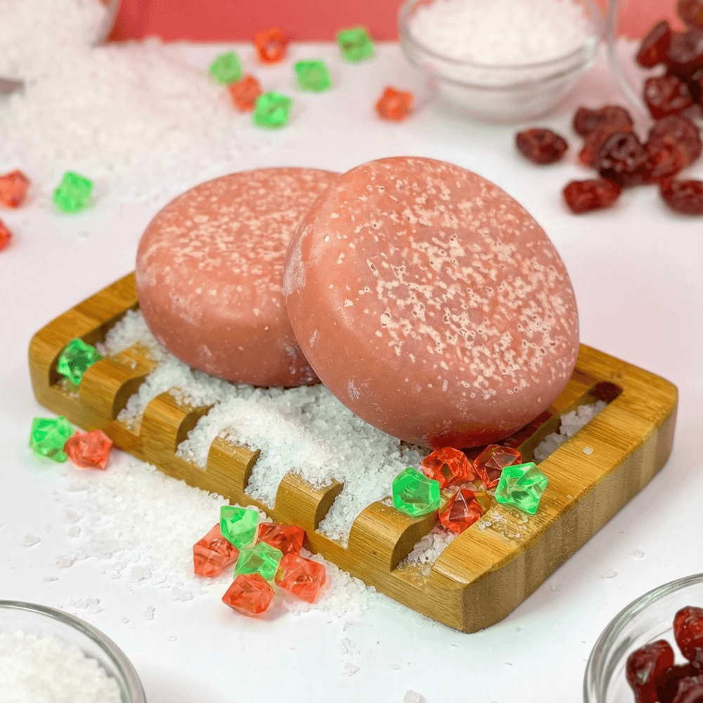 Two coral-pink round bars with white speckling on top, displayed on a bamboo soap dish with white sea salt. Decorative red and green acrylic gems scattered around.