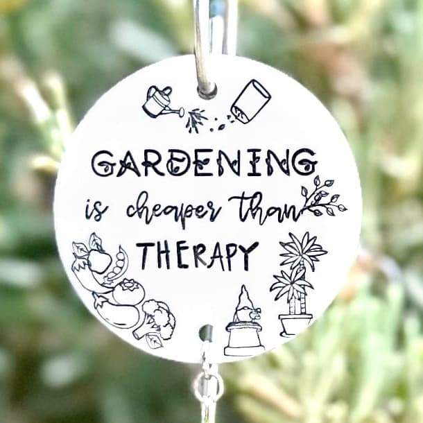 Round white aluminum disc hanging from a metal stake, hand-stamped with text and garden illustrations including watering cans, flowers, and a gnome. Photographed outdoors against blurred greenery.