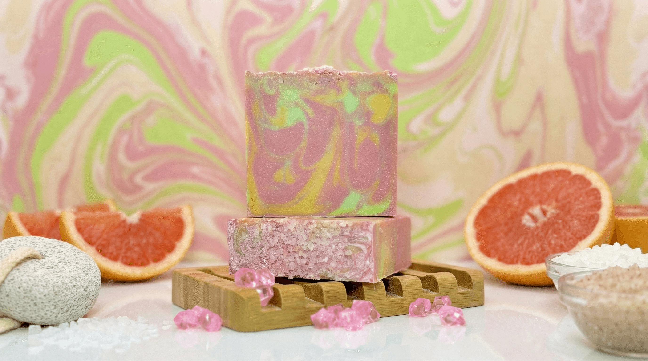 Two stacked handmade soap bars with pink, yellow, and green marbled swirls displayed on a wooden soap dish, surrounded by grapefruit halves, sea salt, pumice stone, and pink salt crystals.