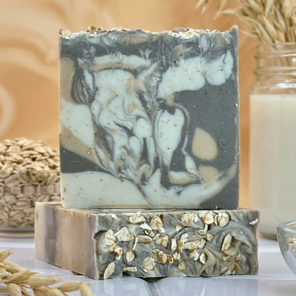 Two stacked bars with marbled gray and cream swirls. The bottom bar is topped with whole oat flakes. Oats, a glass of milk, and rosemary visible in the background.