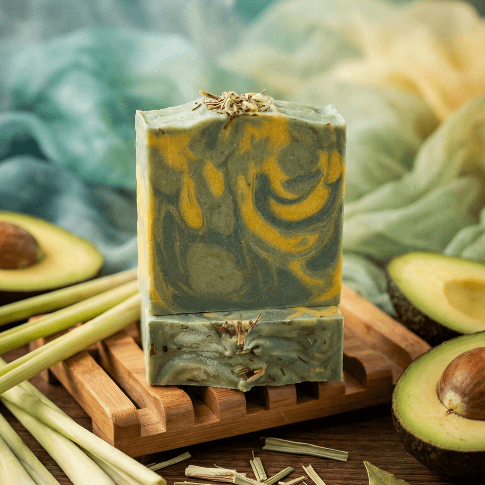 Two stacked bars of swirled olive green and golden yellow soap topped with dried lemongrass pieces, displayed on a wooden soap dish surrounded by fresh lemongrass stalks and halved avocados.