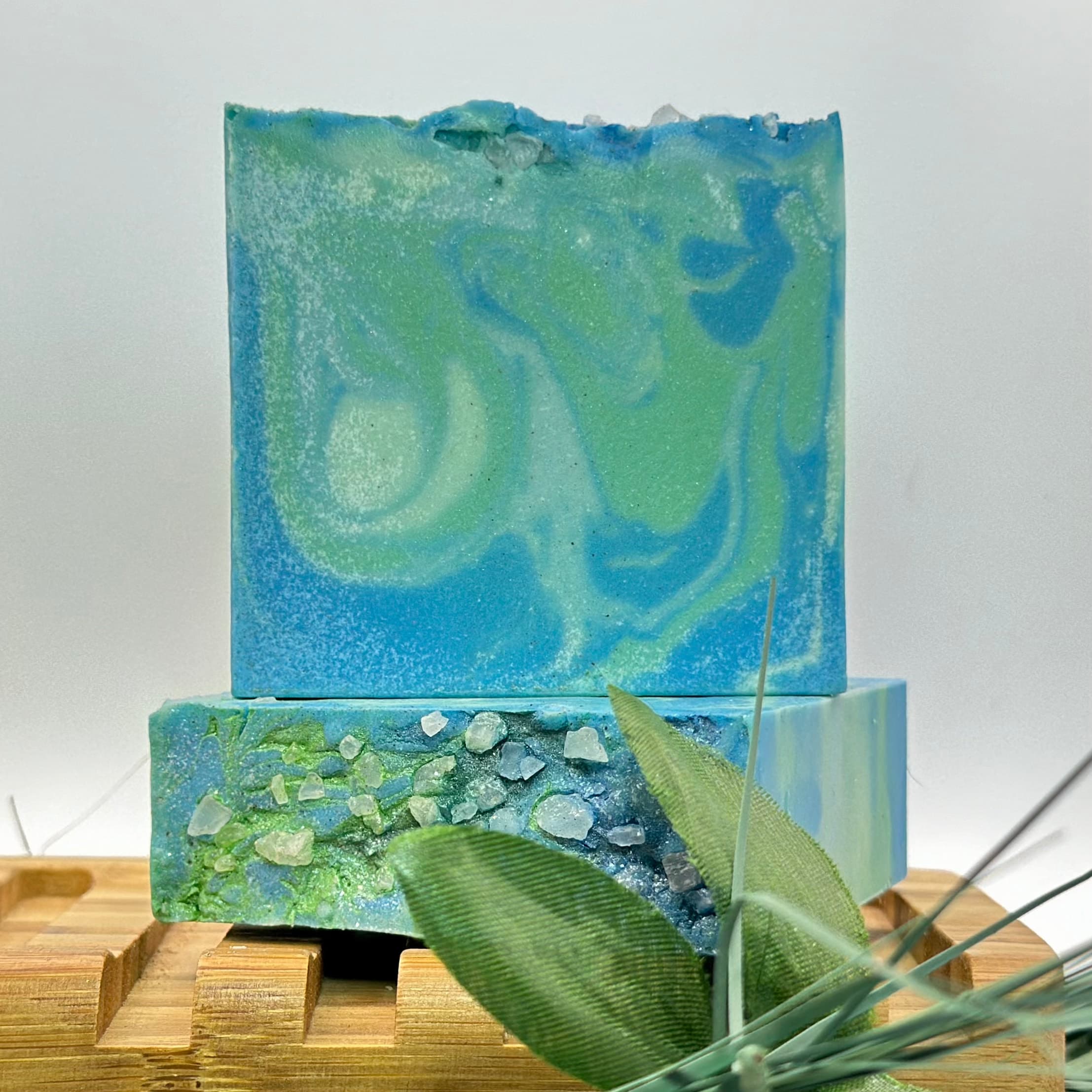 Swirled blue and green soap bars with marbled pattern, displayed on wooden soap deck. Front bar shows embedded sea salt crystals and pumice pieces, with decorative green leaves arranged nearby.