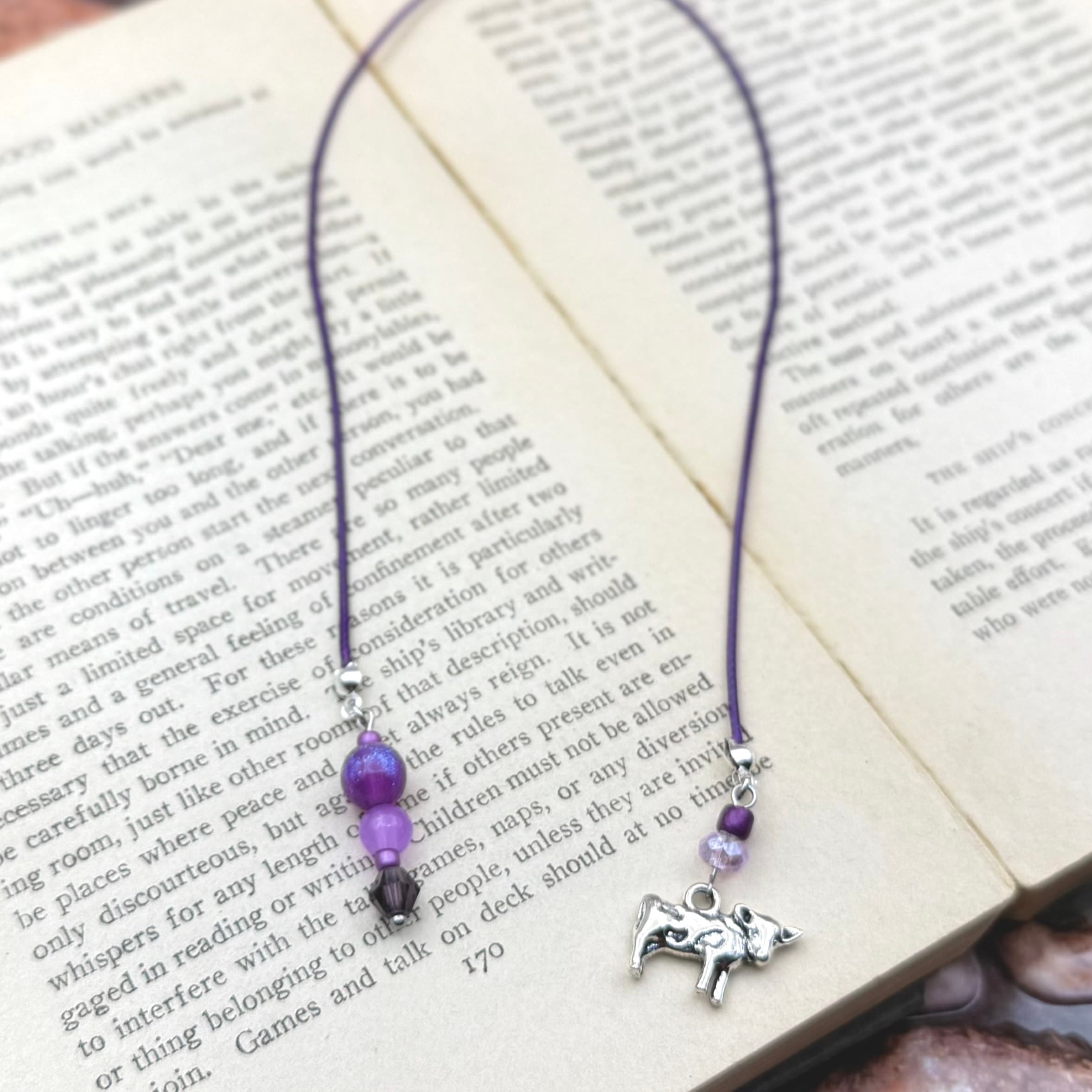 Purple beaded bookmark with silver elephant charm resting on open book pages. Features purple cord with translucent purple and dark accent beads, displayed on aged text.