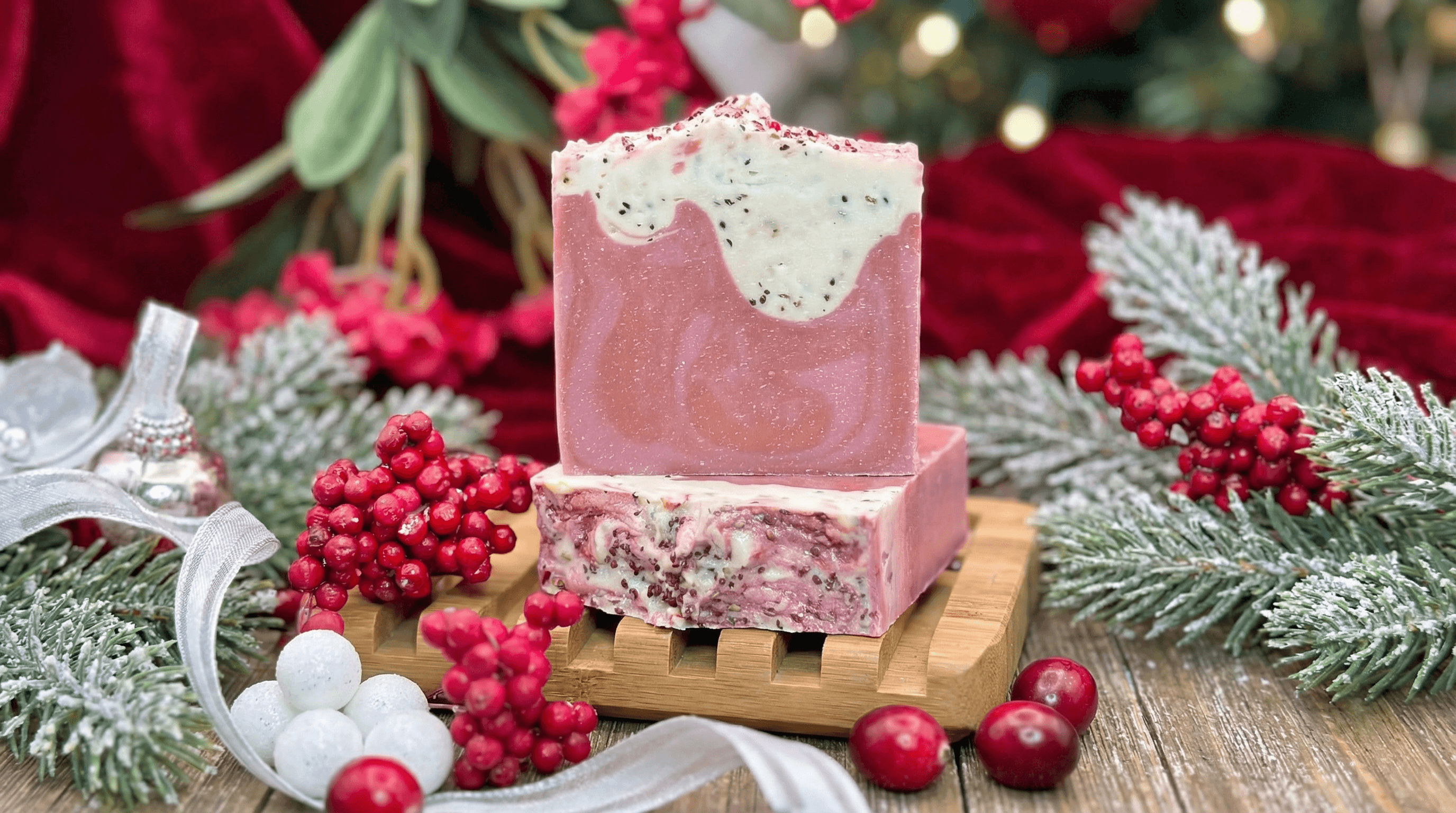 Pink handcrafted soap bar with white frosted top decorated with red and black specks, displayed on wooden soap dish with festive holiday greenery, red berries, and ornaments.