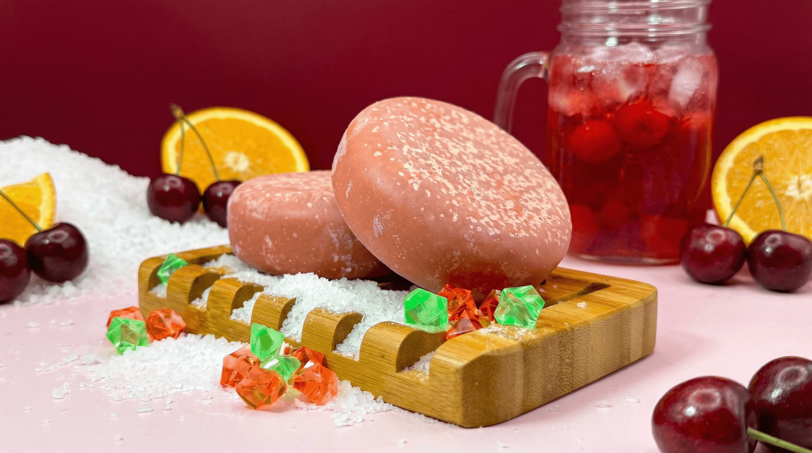 Two coral-pink salt scrub bars with glistening salt crystals on surface, displayed on bamboo tray with white salt, orange slices, cherries, and red drink in mason jar against burgundy background.