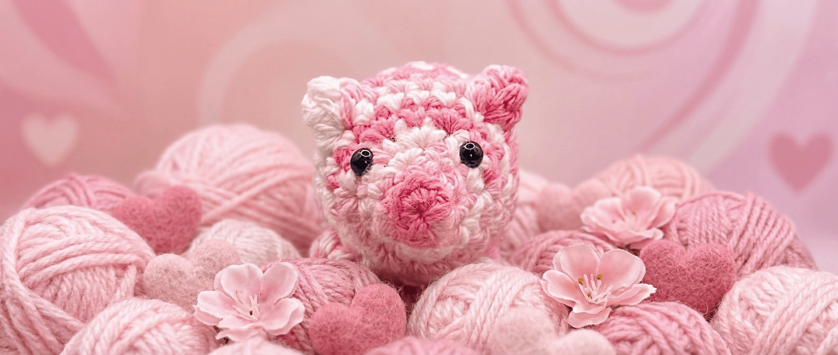 Hand-crocheted pink and white variegated pig sachet with black safety eyes, surrounded by pink yarn balls, pom poms, and delicate pink flowers on a soft pink background.