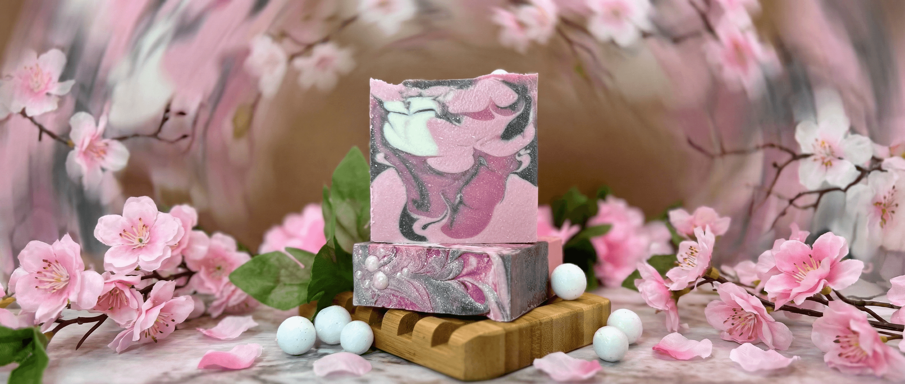 Two bars of marbled pink, gray, and white handcrafted soap stacked on wooden dish, surrounded by pink cherry blossoms and white bath pearls in a soft-focus floral setting.