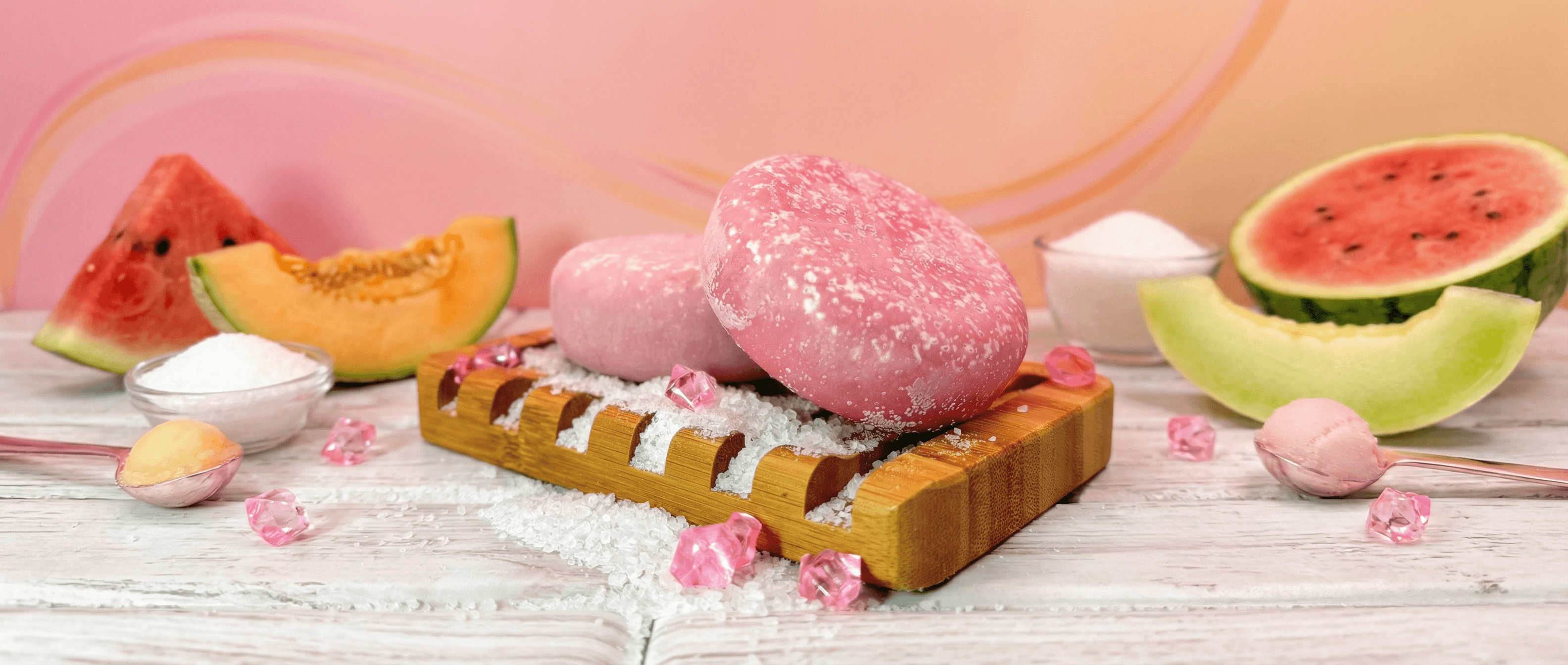 Pink oval sea salt scrub bar with crystallized sugar coating, displayed on wooden soap dish with scattered salt, surrounded by fresh watermelon and cantaloupe slices on white wood surface.