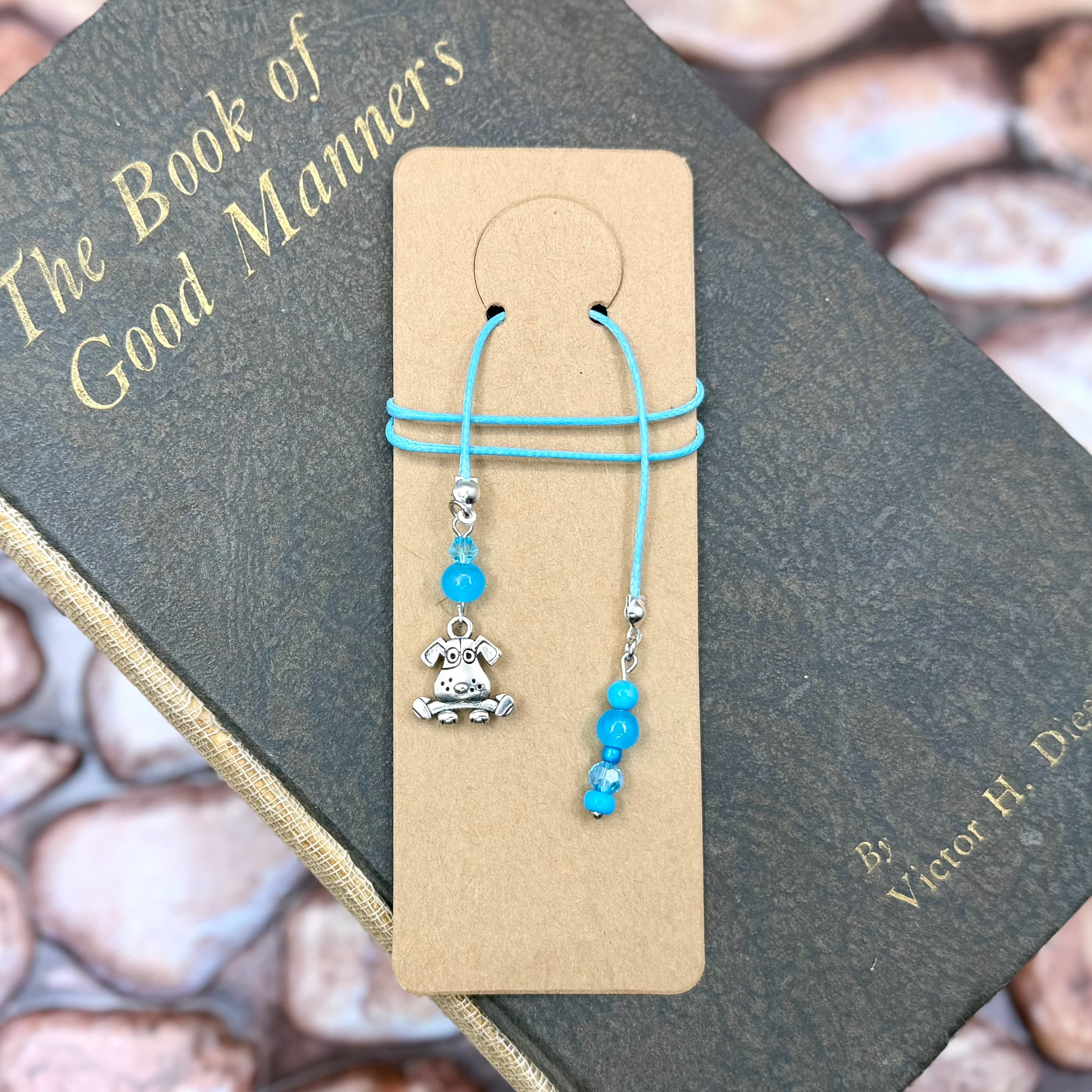 Handmade bookmark with turquoise cord and blue beads features a silver dog charm. Displayed on tan cardstock atop a vintage book titled 'The Book of Good Manners'.