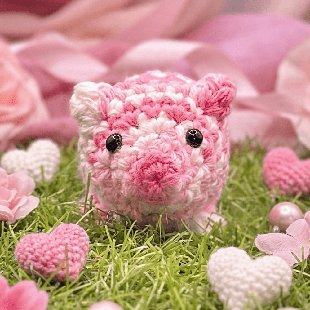 Hand-crocheted pink and white variegated pig sachet with black bead eyes and small ears, sitting on artificial grass surrounded by pink hearts and flower petals.
