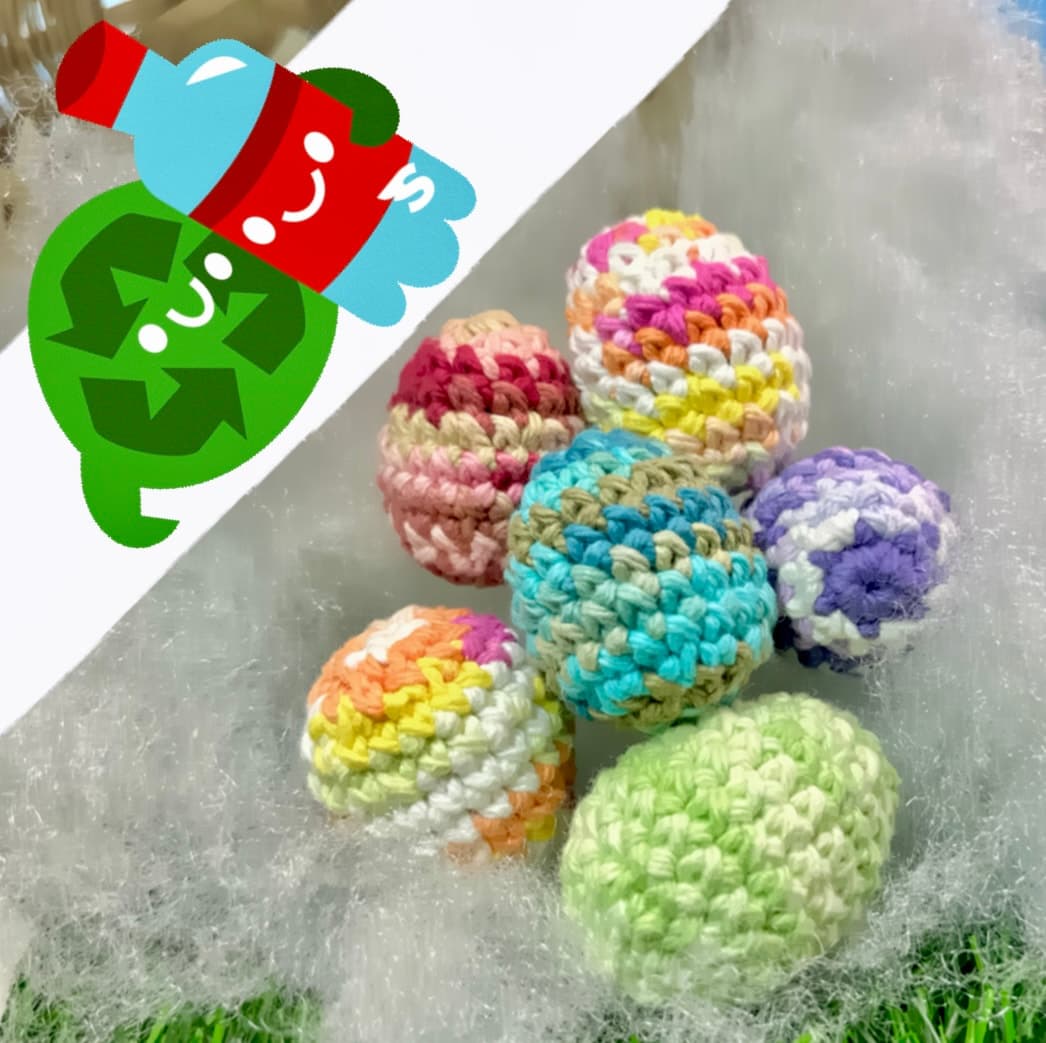 Six colorful crocheted eggs in pastel stripes and variegated patterns, arranged on a gray surface next to a card with cartoon recycling characters. Each egg features unique color combinations.