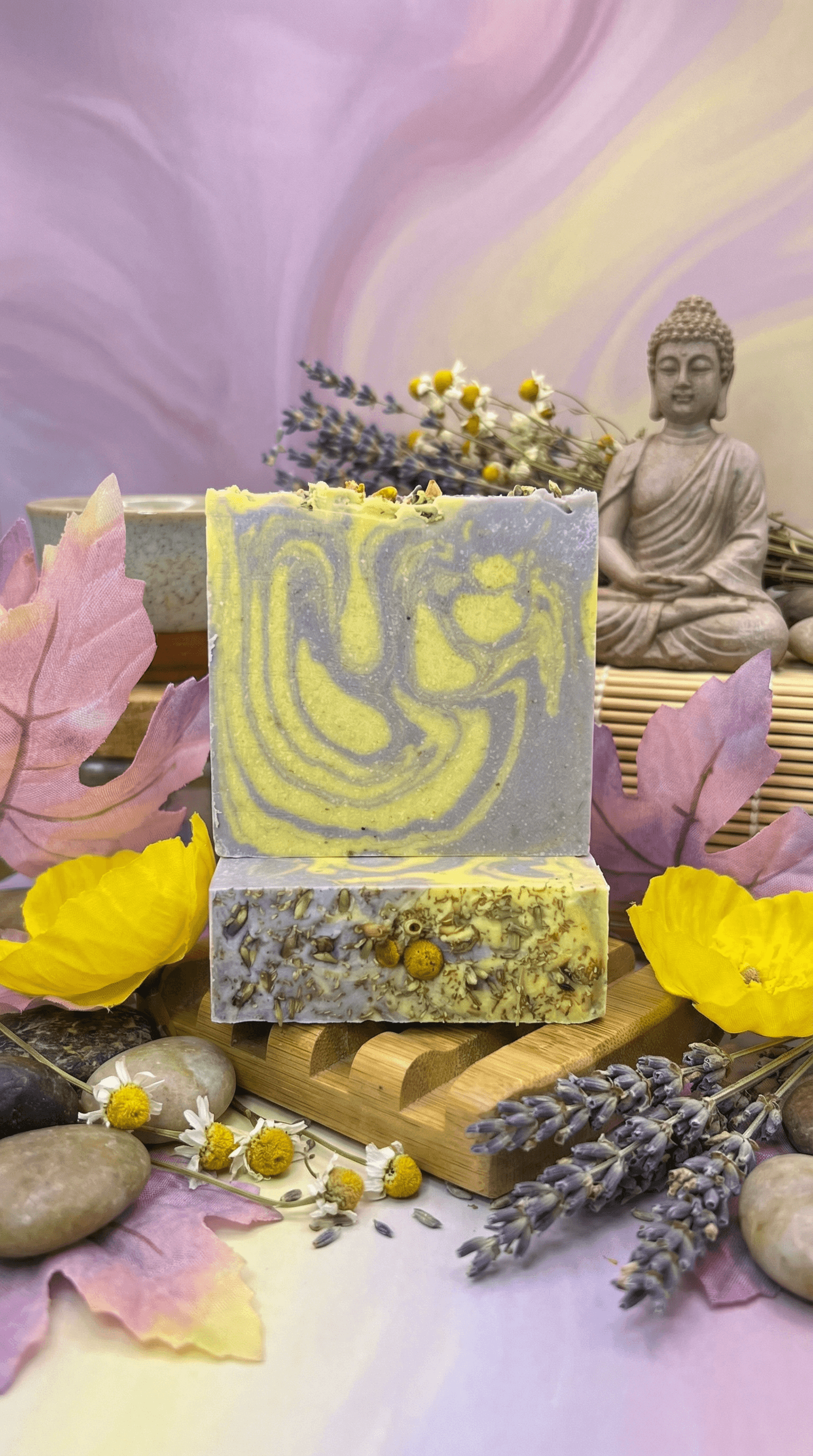 Two stacked handmade soap bars with yellow and purple swirled top, chamomile-studded base with dried lavender buds, displayed on bamboo tray with Buddha statue, stones, and botanicals.
