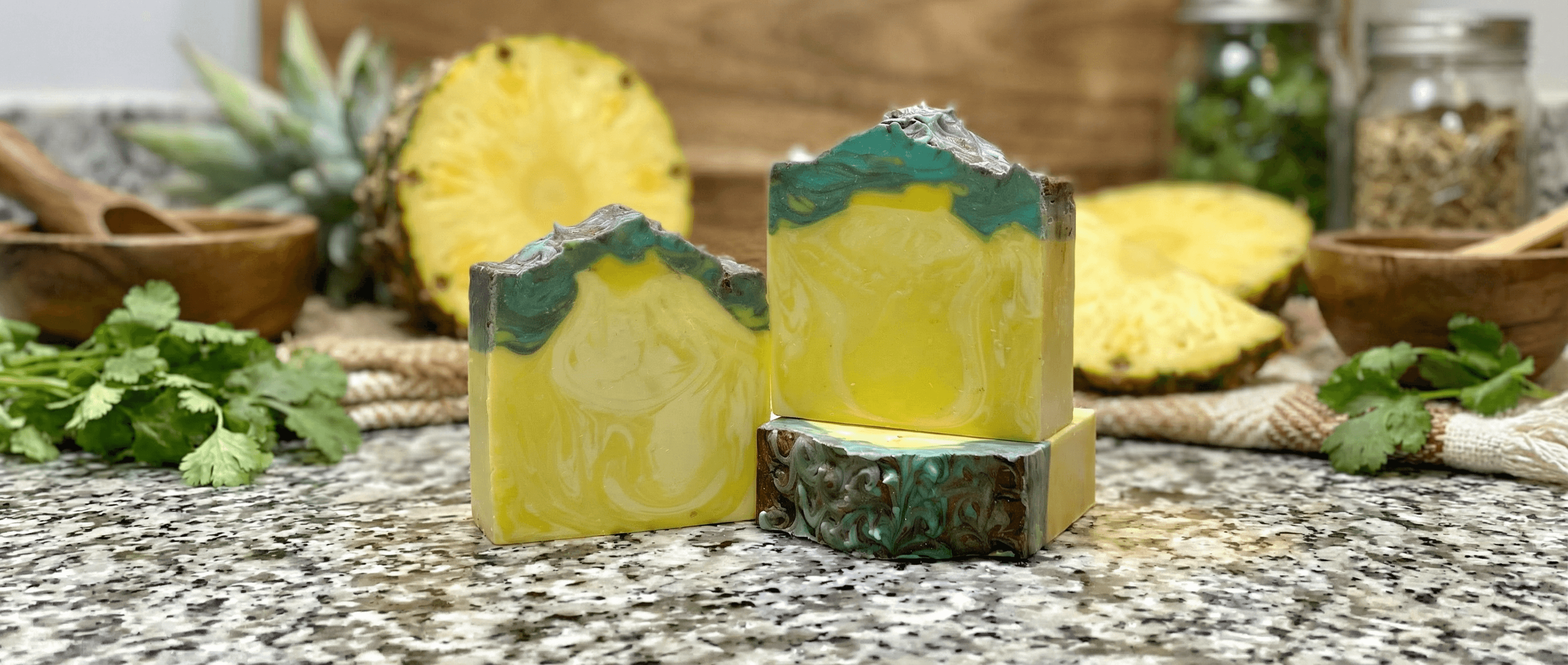 Two bars of bright yellow handcrafted soap with green and gray swirled tops, displayed on granite with fresh pineapple, cilantro, and wooden bowls in the background.