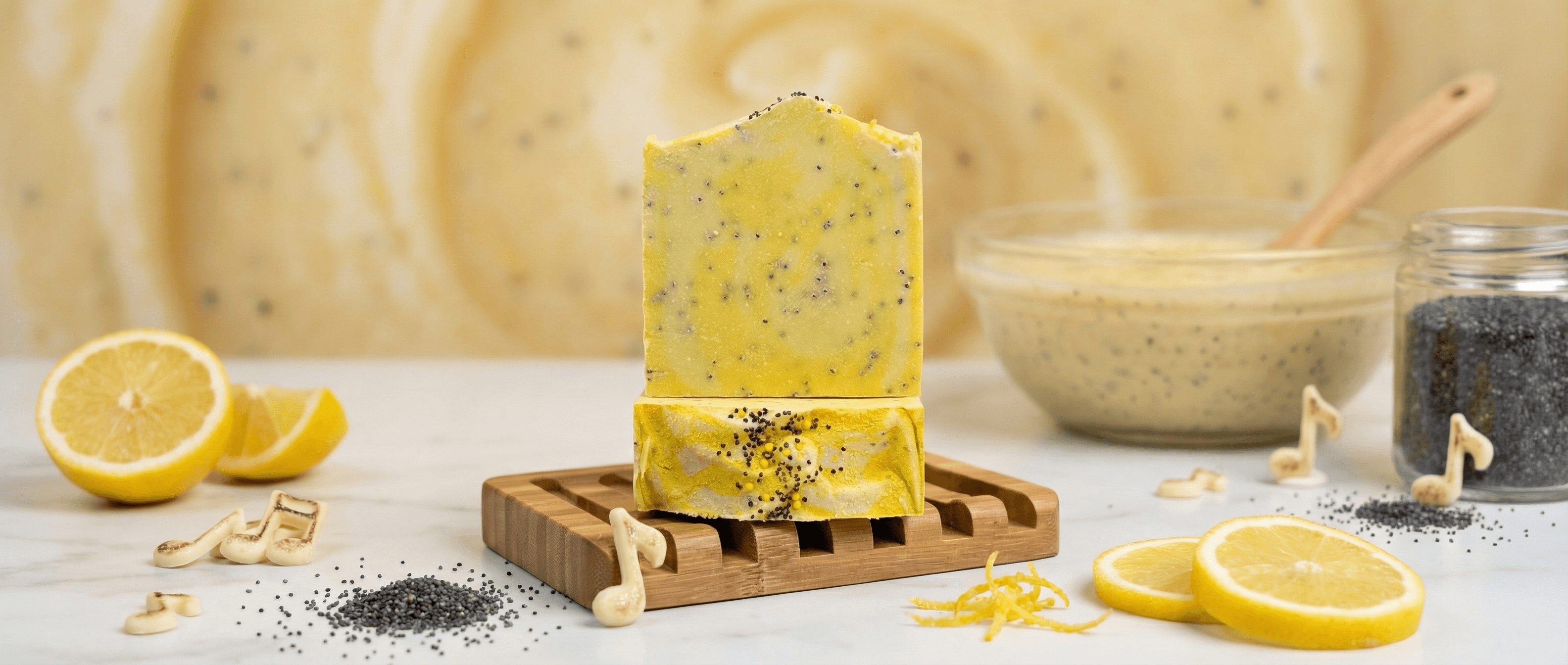 Bright yellow handcrafted soap bars with black poppy seeds throughout, stacked on a wooden soap dish. Fresh lemon slices and loose poppy seeds arranged around the display.