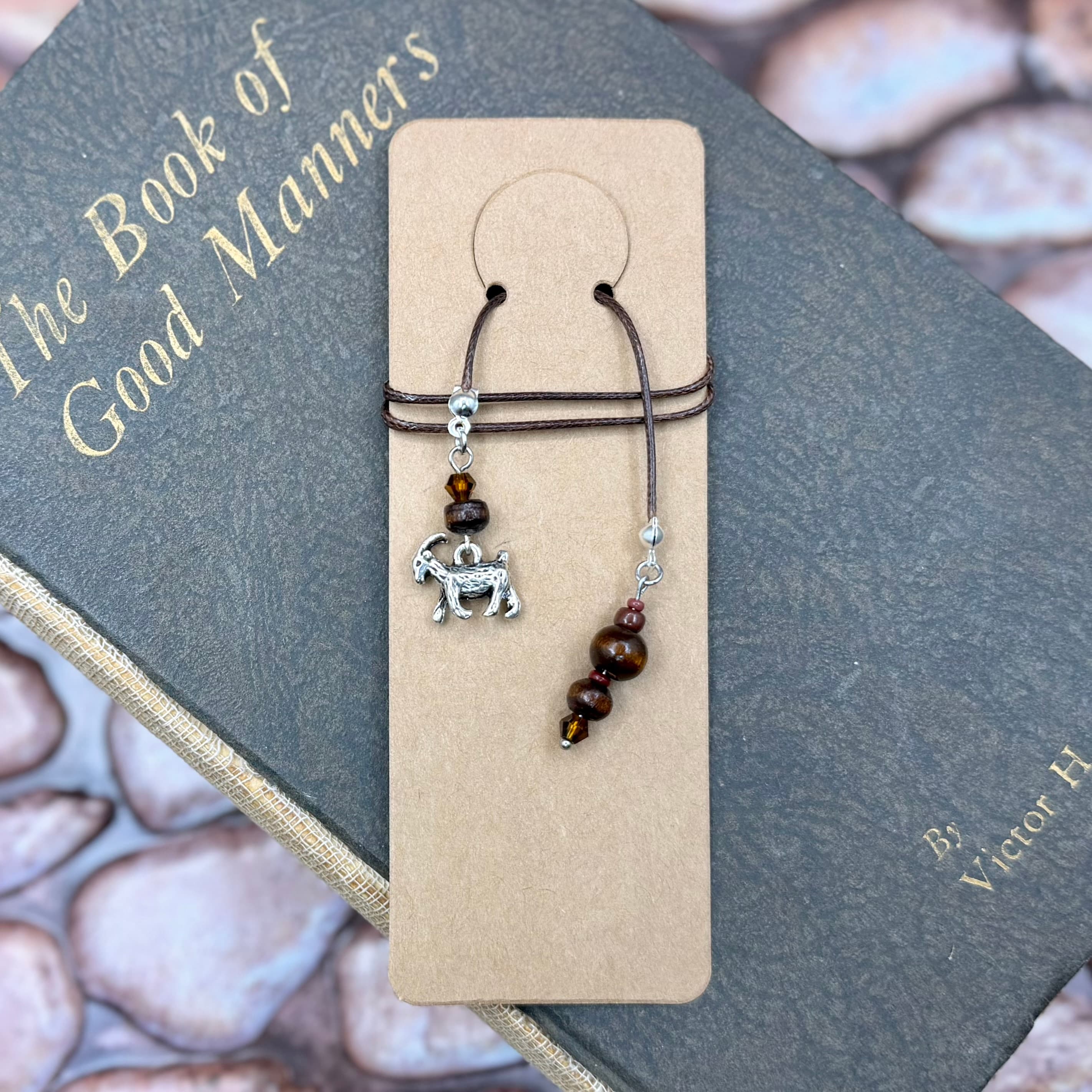 Handmade brown goat charm bookmark with wooden beads on brown cord, displayed on tan card over a vintage book titled 'The Book of Good Manners' with lace fabric visible.