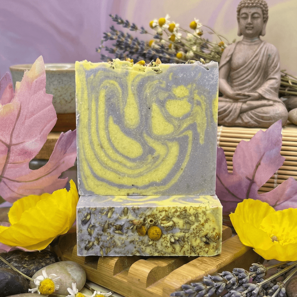 Two stacked handmade soap bars with purple and yellow swirled top, dried chamomile and lavender bottom layer, displayed with Buddha statue, pink and yellow flowers, and dried botanicals.