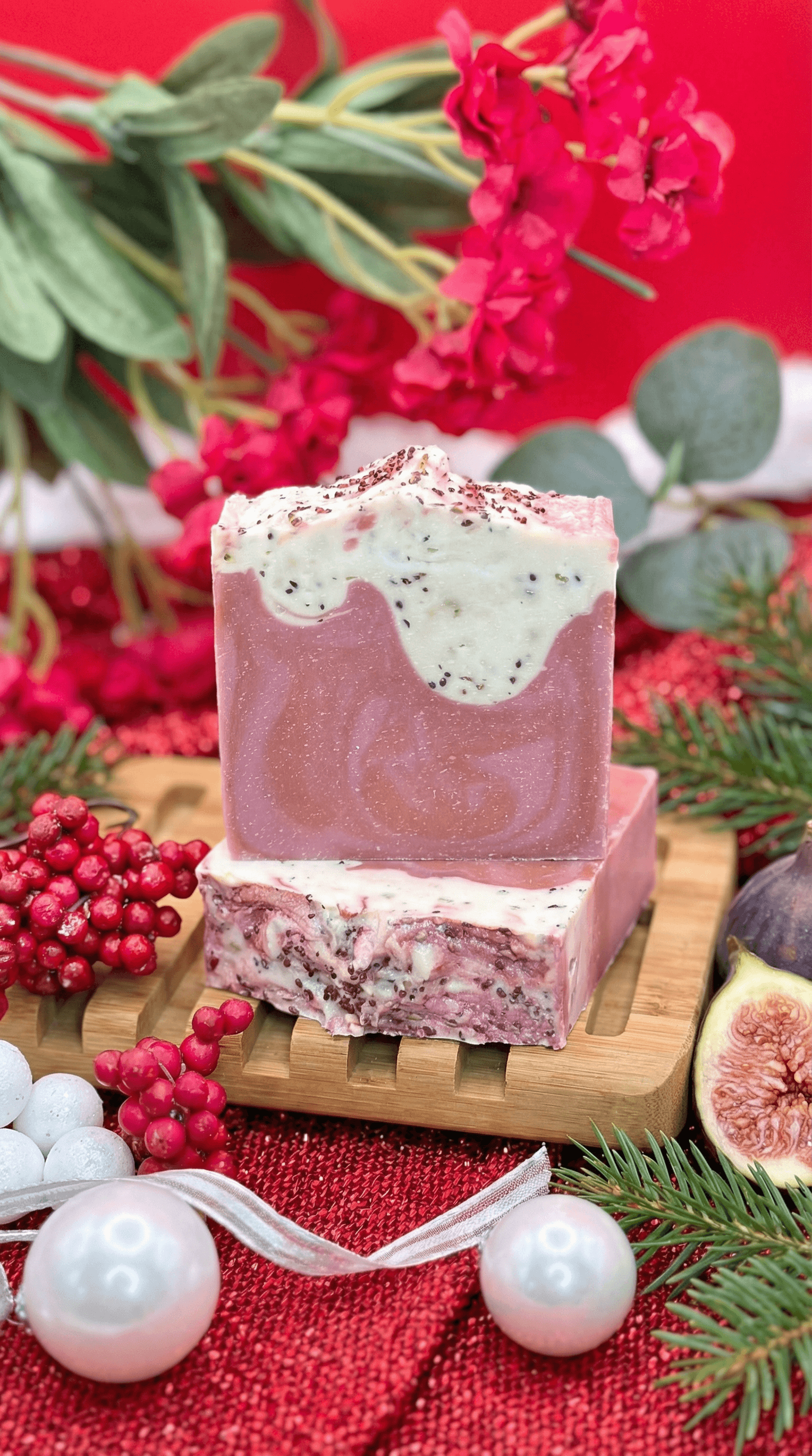 Two stacked bars of handmade soap in dusty rose with white drizzled frosting tops, speckled with dark seeds. Displayed on wooden soap dish surrounded by fresh figs, cranberries, and winter greenery.