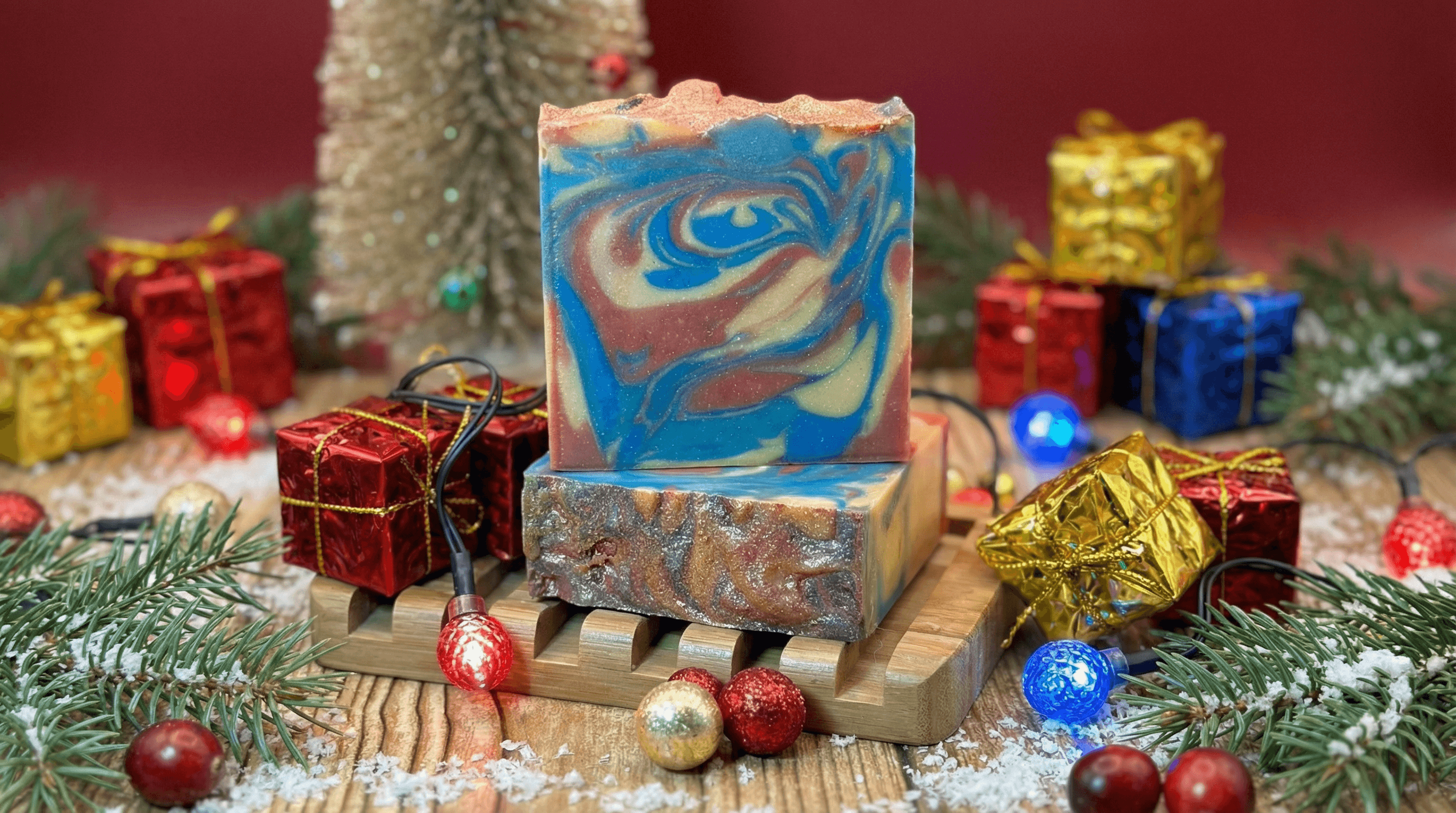 Swirled handmade soap bar in blue, burgundy, and cream with glittery gold top, displayed on wooden soap dish surrounded by miniature wrapped gift boxes and pine branches.