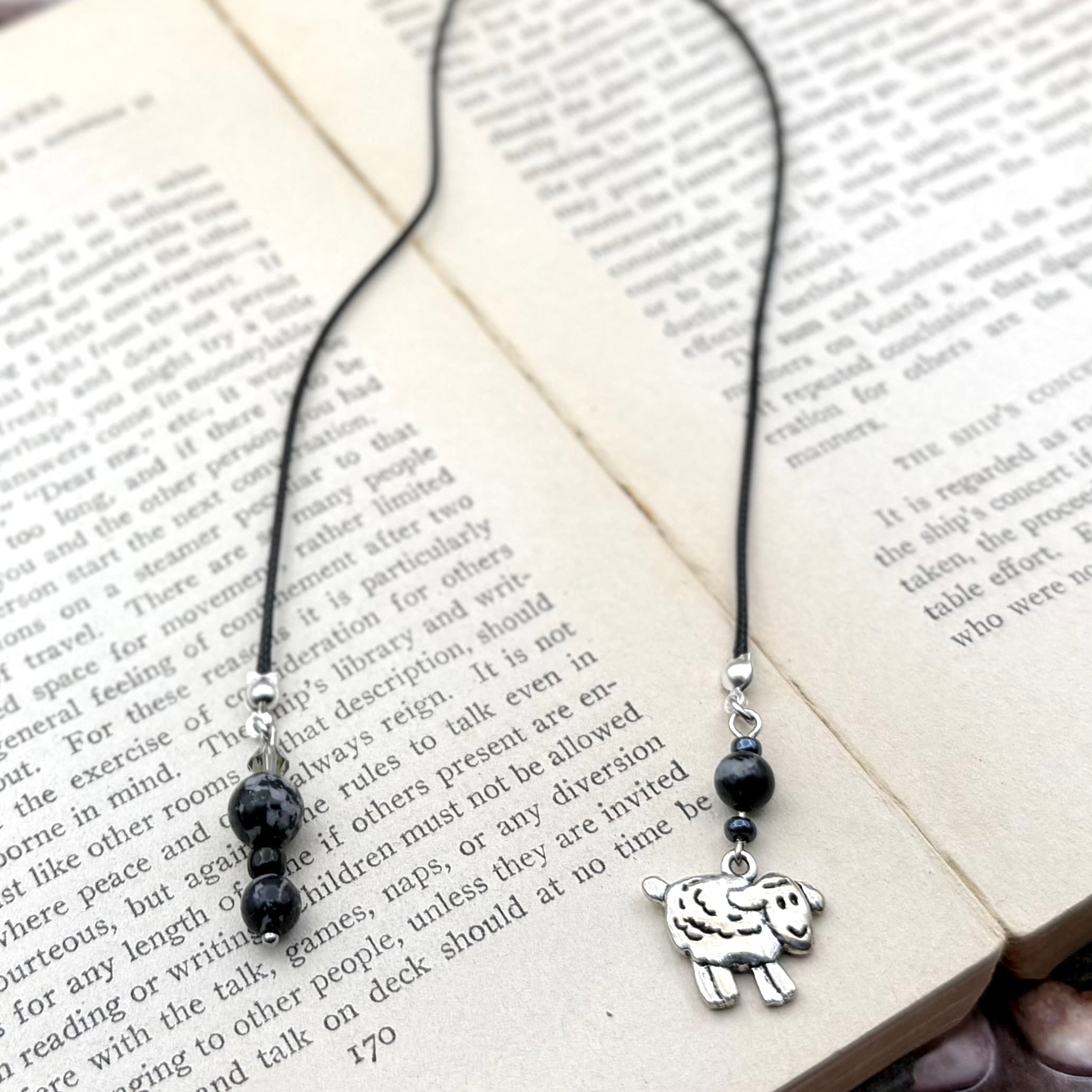 Handmade bookmark with black cord and speckled black beads, featuring a silver-toned sheep charm, displayed on open book pages with vintage text.