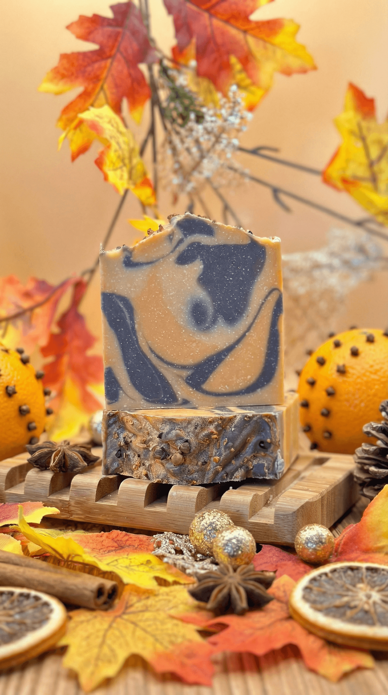 Handcrafted soap bar with orange and navy blue swirls on top, textured bottom layer with embedded cloves and botanicals, displayed on wooden soap dish surrounded by autumn leaves, dried orange slices, cinnamon sticks, and star anise.
