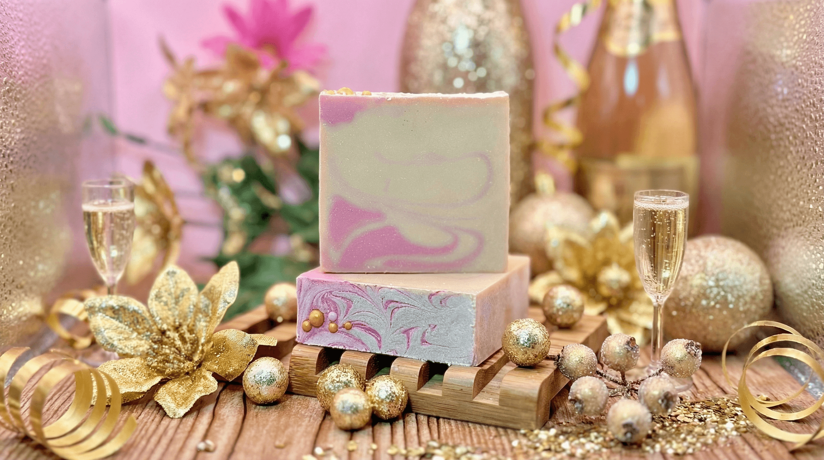 Two bars of marbled pink and cream soap with swirled patterns, displayed on a wooden soap dish surrounded by gold holiday decorations, champagne flutes, and glittery ornaments.