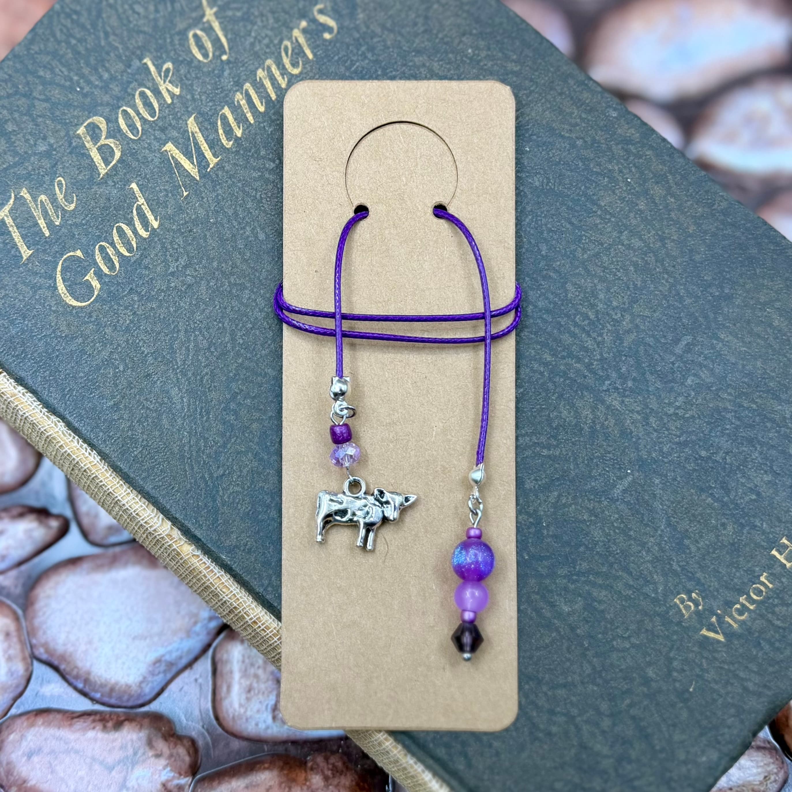 Handmade bookmark with purple cord, silver cow charm, and purple beaded tassel displayed on tan card, resting on vintage book titled 'The Book of Good Manners'.