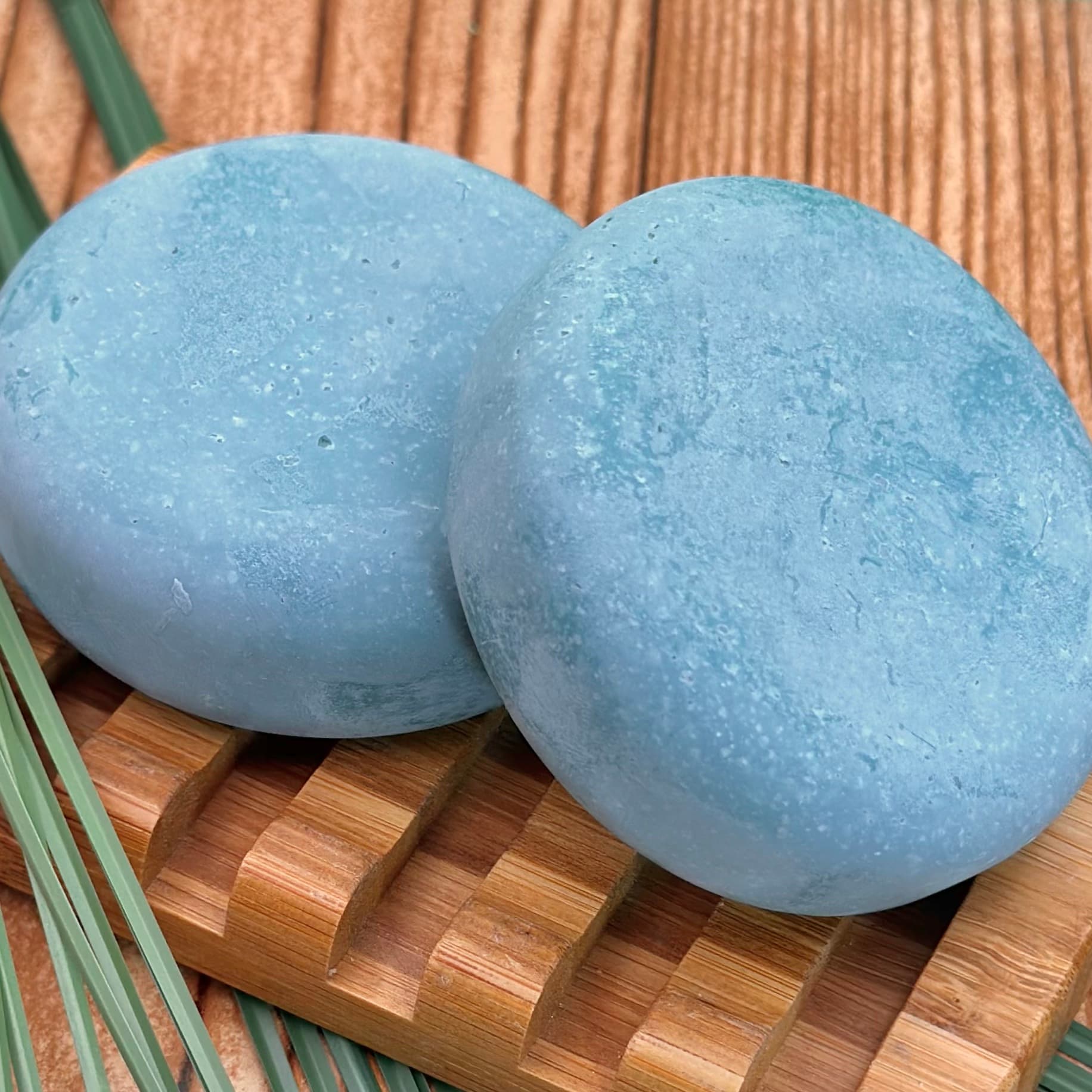 Two pale blue round soap bars with visible sea salt crystals on surface, resting on a woven bamboo soap dish with green palm leaves in background.