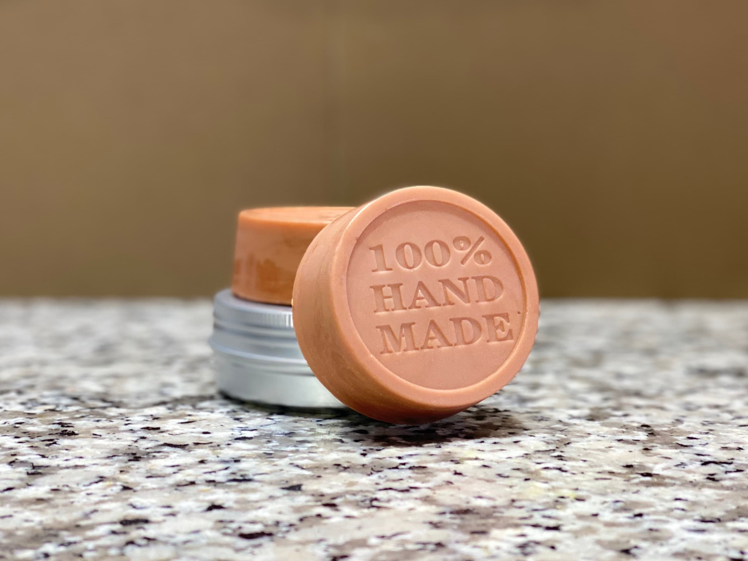 Peach-colored round solid lotion bar with embossed '100% HAND MADE' text, displayed with silver tin and additional bar on speckled granite surface against warm tan background.