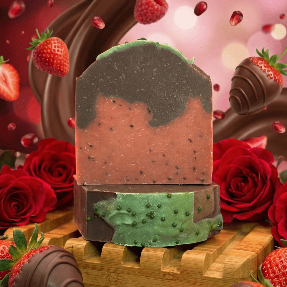 Two stacked handcrafted soap bars on bamboo dish. Top bar is coral-pink with dark speckles and green top layer. Bottom bar is translucent green with suspended bubbles. Surrounded by red roses, strawberries, and chocolate-dipped berries.