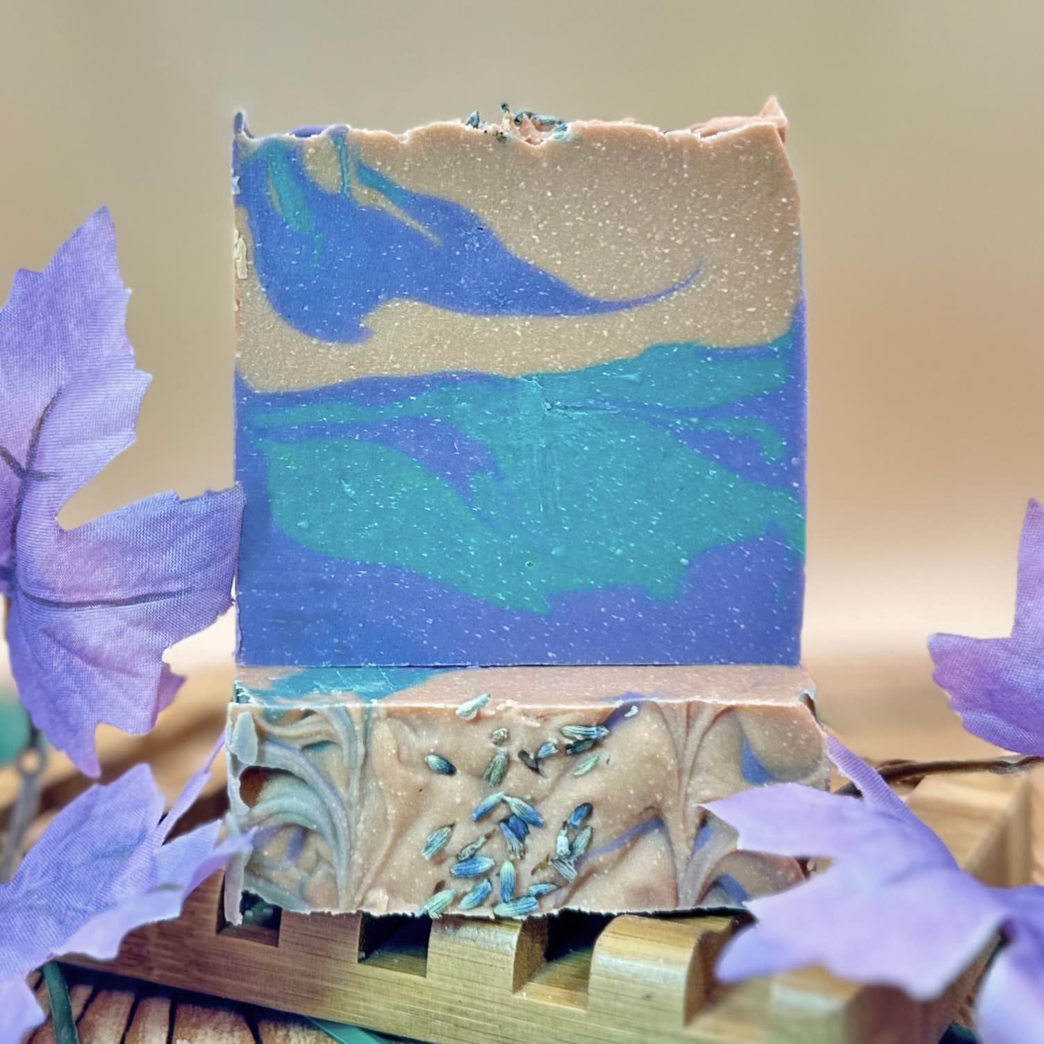 Handcrafted soap bar with swirled layers of blue, turquoise, and tan colors with sparkly texture. Dried lavender buds are sprinkled on top and bottom, displayed on a wooden soap dish.