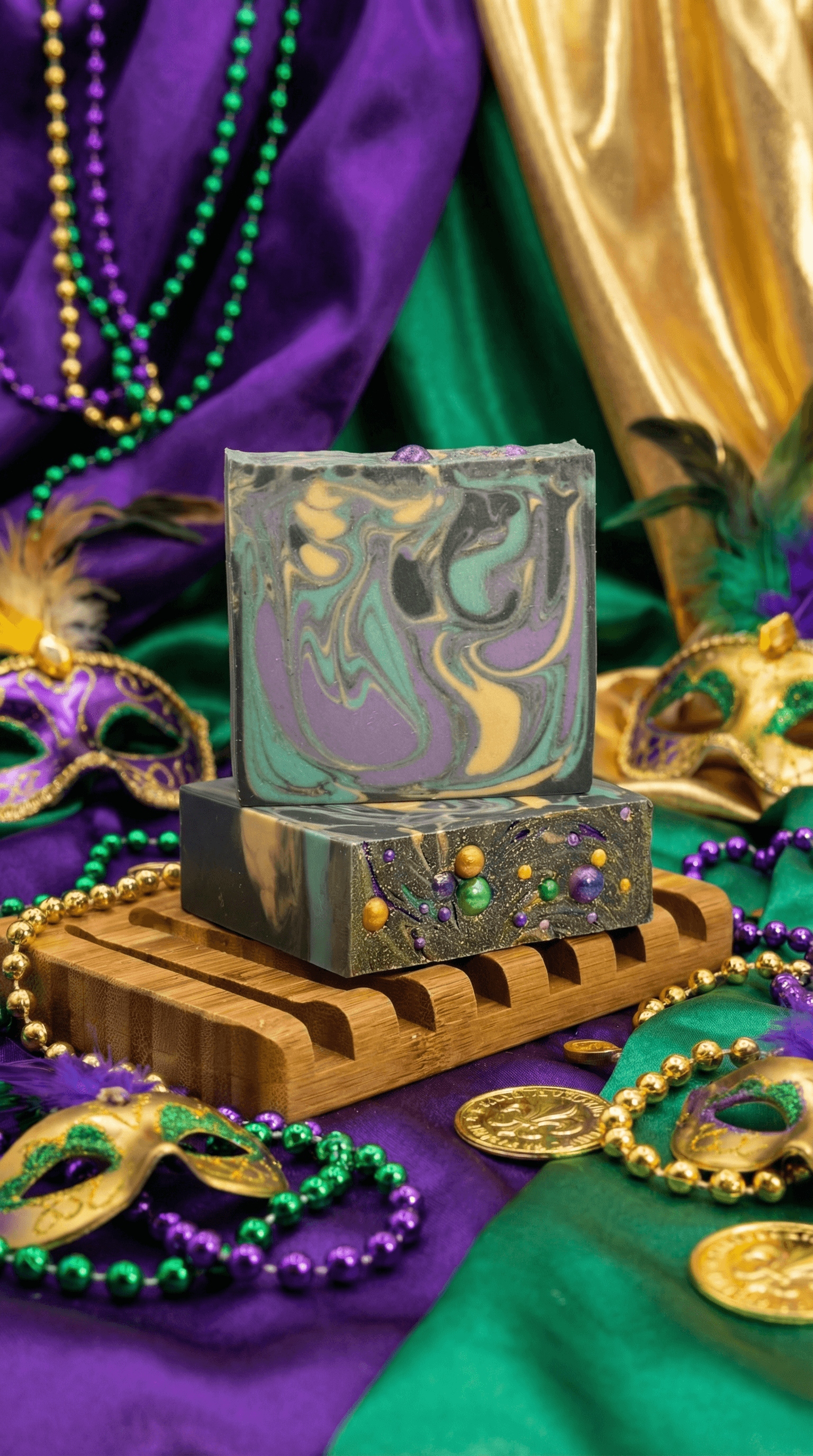Two marbled soap bars in purple, green, gold, and black swirls stacked on a bamboo soap dish, surrounded by Mardi Gras beads, masks, coins, and fabric in traditional carnival colors.