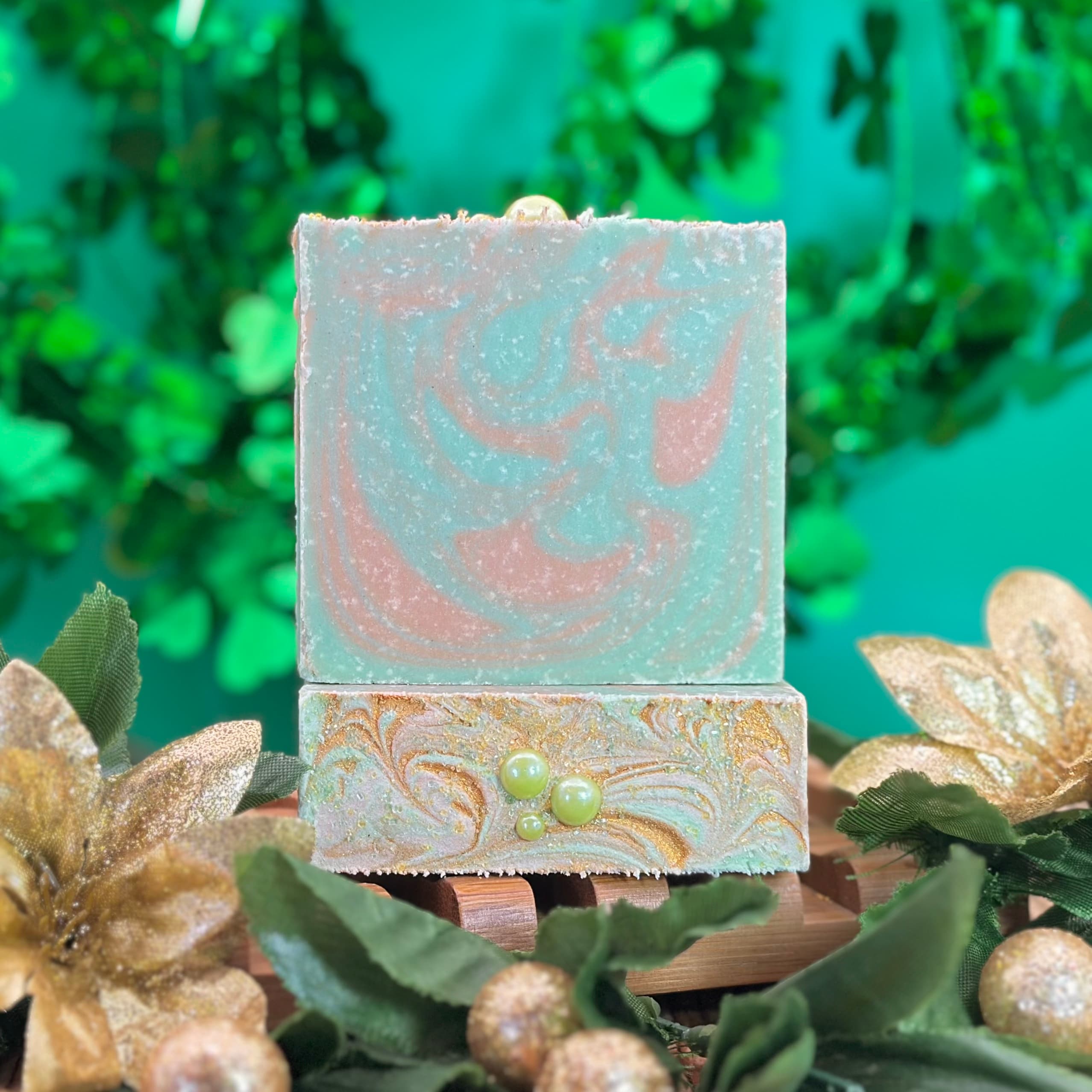 Two stacked handcrafted soap bars with swirled teal and rose gold marbling. The front bar features gold shimmer swirls and small green pearl embeds, displayed on a wooden soap dish with gold botanical accents.