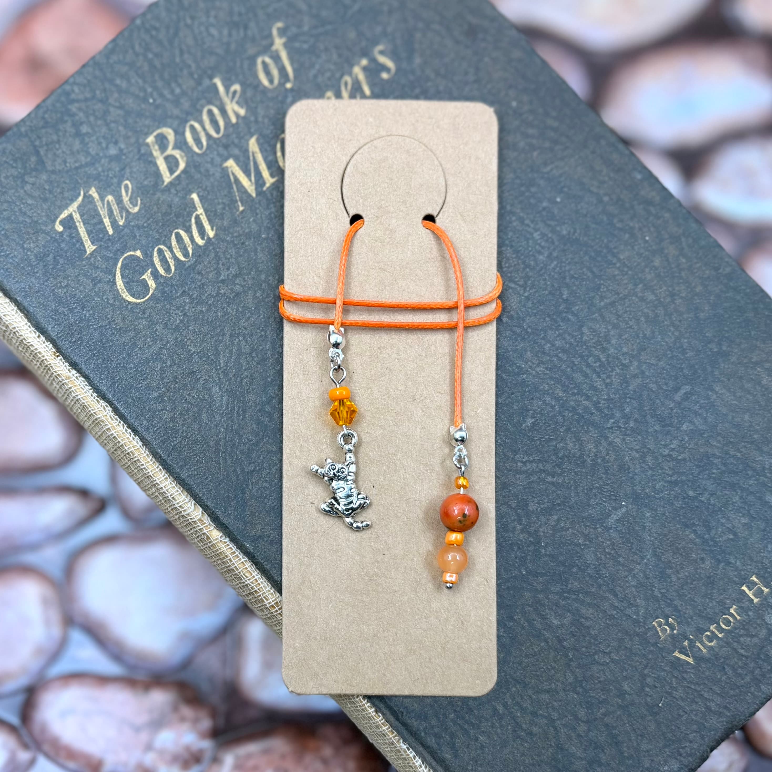 Handmade bookmark with orange cord, silver cat charm, and amber beads displayed on tan card stock atop a vintage book titled 'The Book of Good Manners' by Victor H.