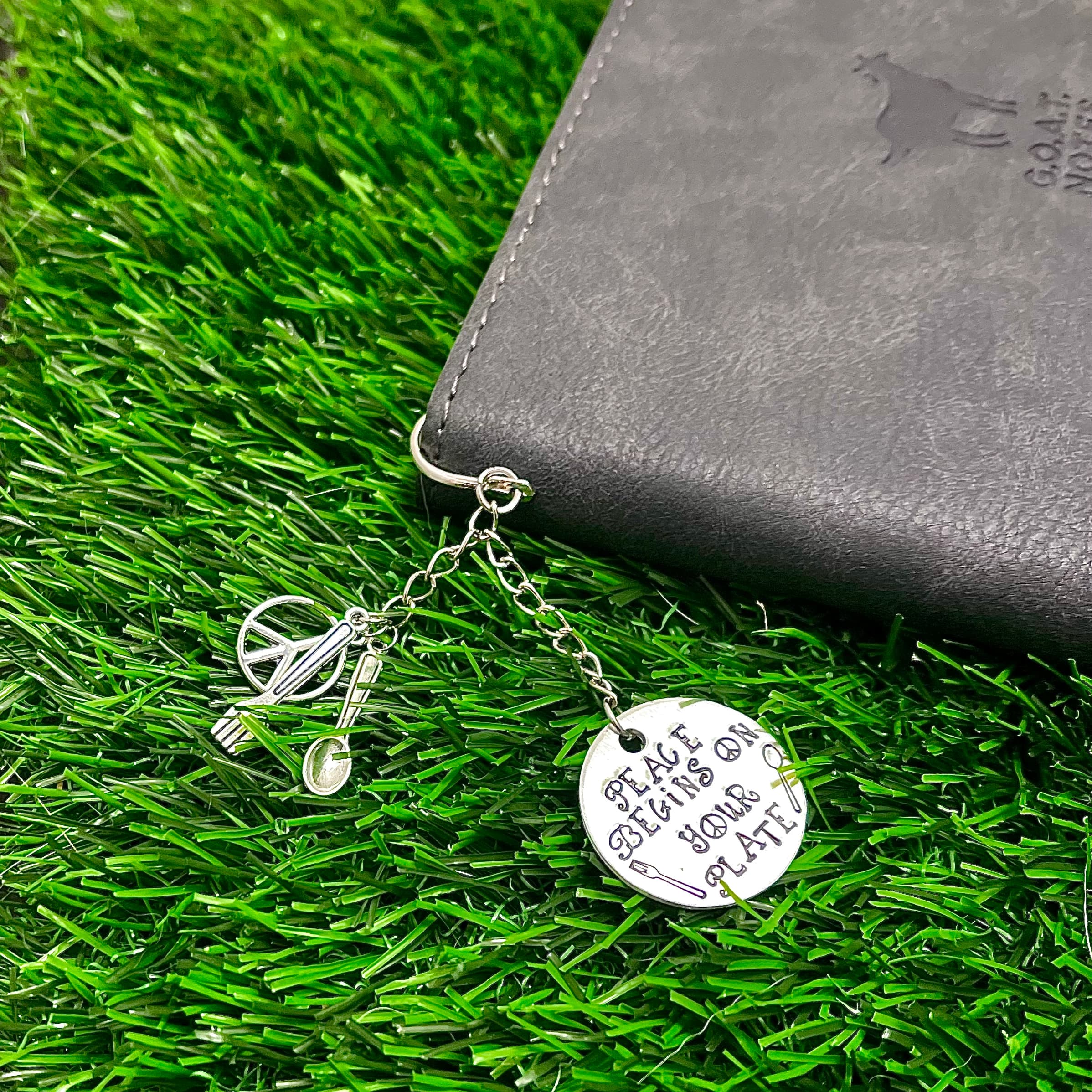 Silver keychain with round stamped disc reading 'Peace Begins On Your Plate' and decorative fork charm, displayed on green artificial grass beside grey vegan wallet.