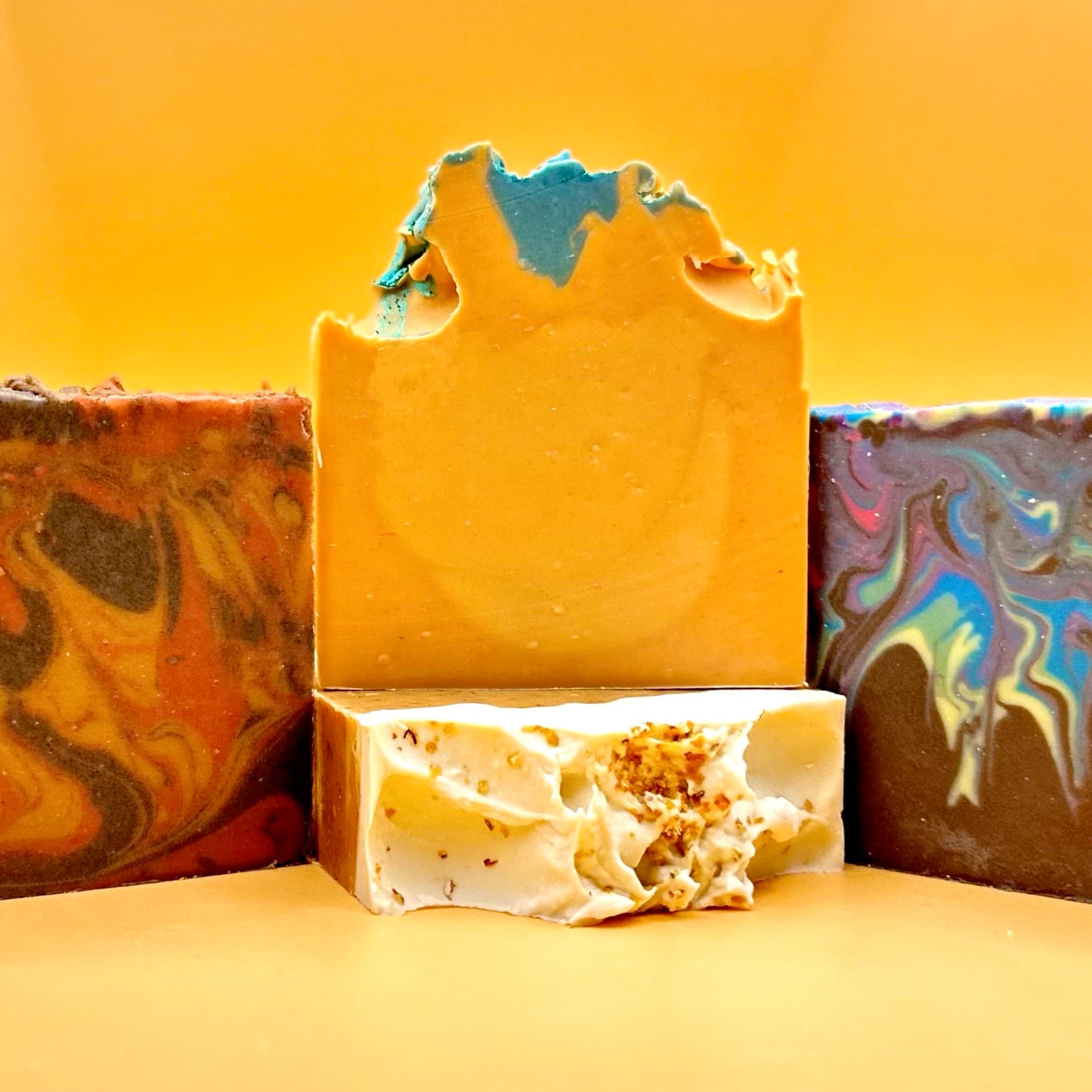 Four handcrafted soap bars in warm autumn tones: swirled brown and olive, solid peachy-orange with blue accent on top, white with herb speckles, and iridescent purple-blue marbling.