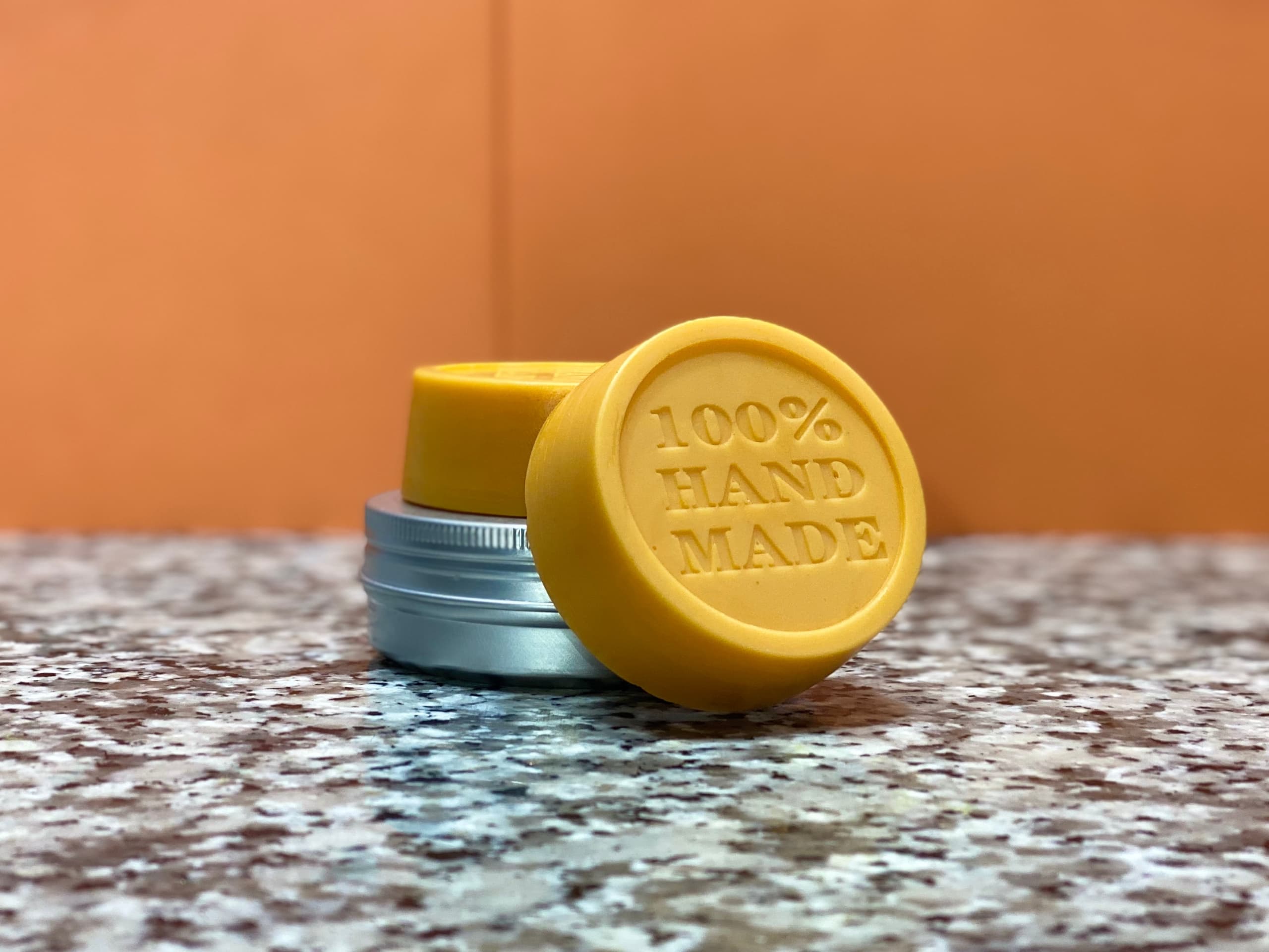 Two round yellow solid lotion bars with embossed '100% HAND MADE' text, displayed on a speckled surface with a small metal tin. One bar stands upright showing the stamped design.