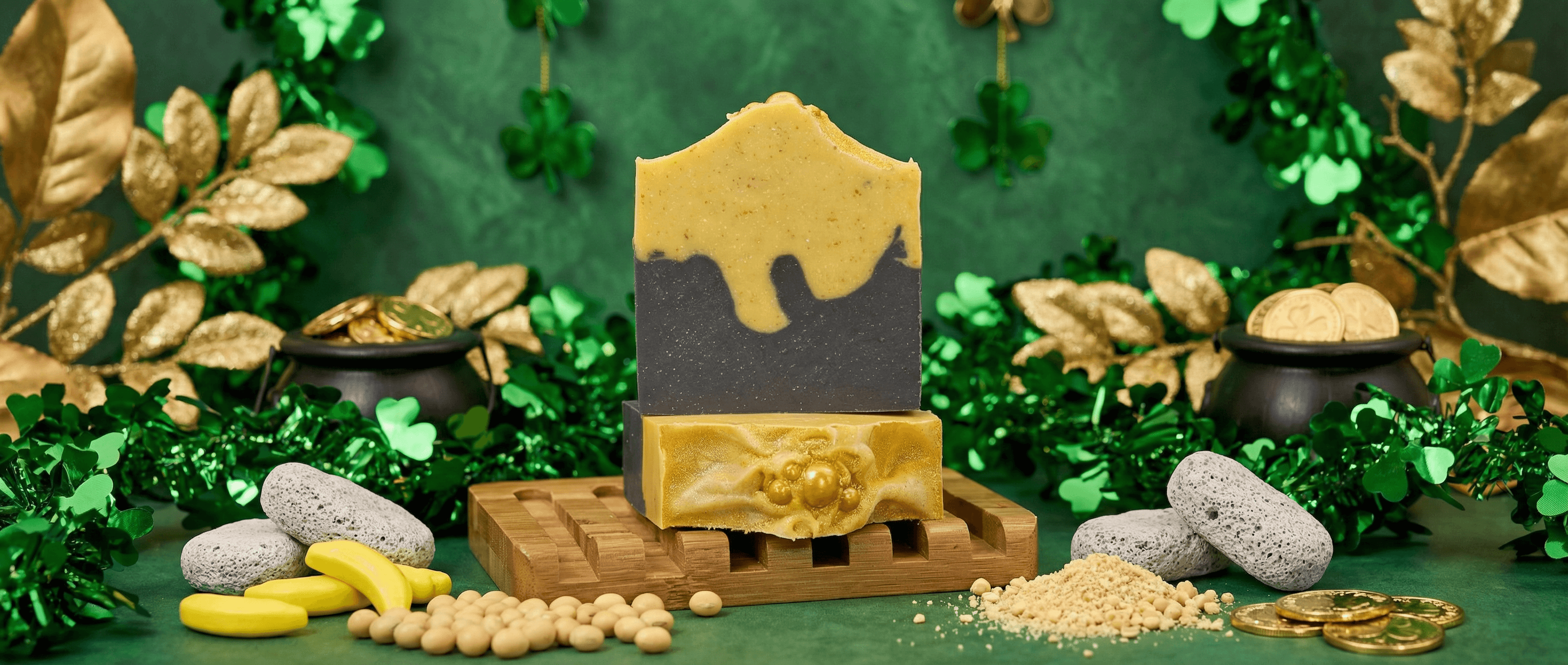 Two-toned handcrafted soap bar with charcoal gray bottom and golden yellow top separated by wavy drip design, displayed on wooden soap dish against festive green backdrop with gold coins and shamrock decorations.