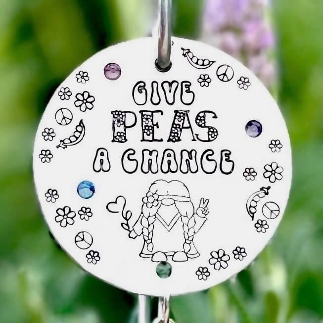 Round white aluminum disc with hand-stamped text and hippie gnome illustration, decorated with colorful glass beads in purple, blue and green. Mounted on metal garden stake against blurred outdoor background.